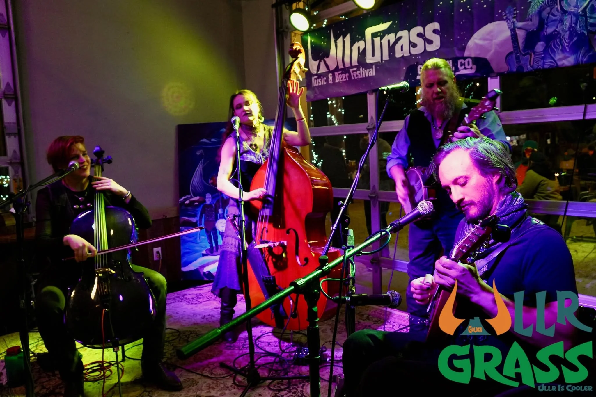 A group of four musicians performing at the Ulrurass Music & Beer Festival. They are playing string instruments, including a cello and guitars, on a stage illuminated by colorful lights with a banner in the background.