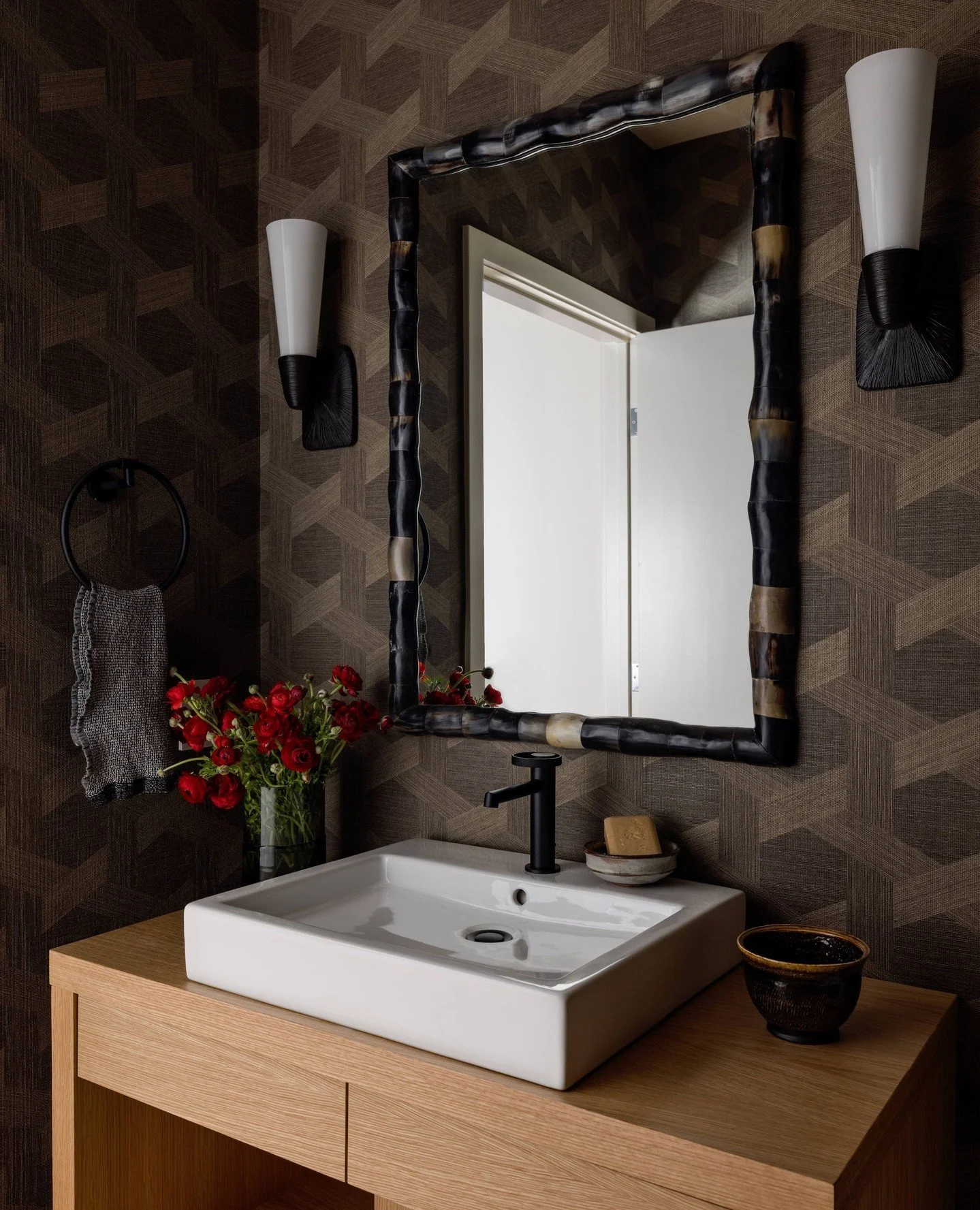We dramatically transformed the functional powder room of our Seattle Skyline Project, with details such as a geometric wall covering, a horn-rimmed mirror, bronze sconces, and a modern wood vanity with a striking Italian faucet. This moody room cont