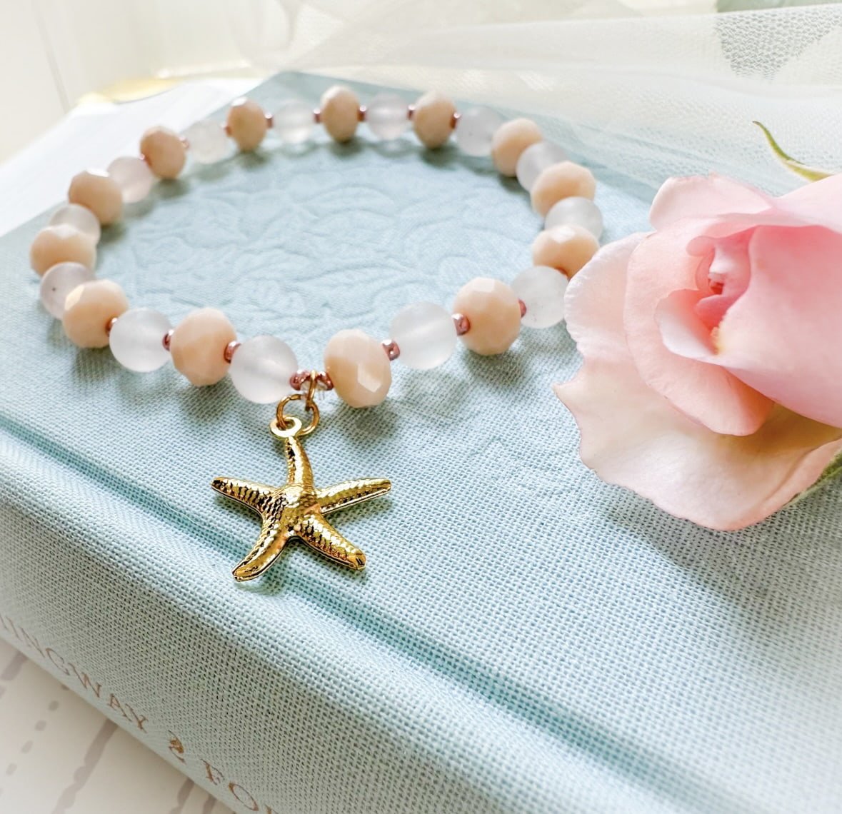 A rose quartz and ivory milk glass elastic bracelet with gold tone starfish charm. Next to a pink rose.