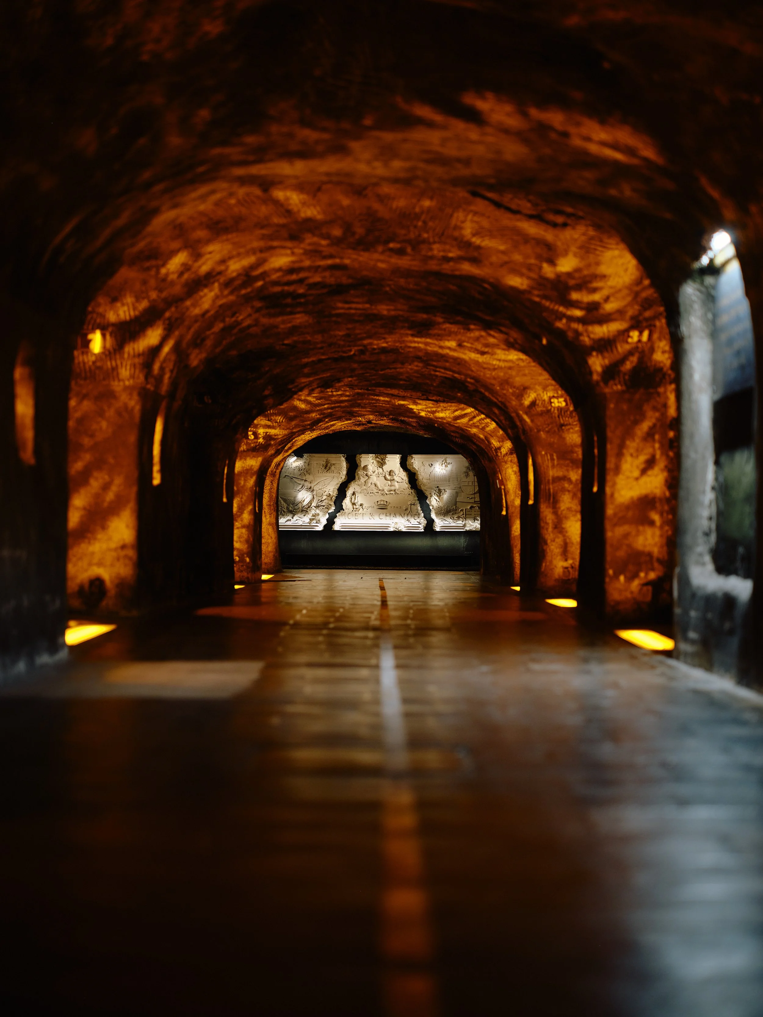 An underground tunnel with warm orange lighting on the ceiling, with a carved sculpture at the end that appears to be a relief of figures and text.