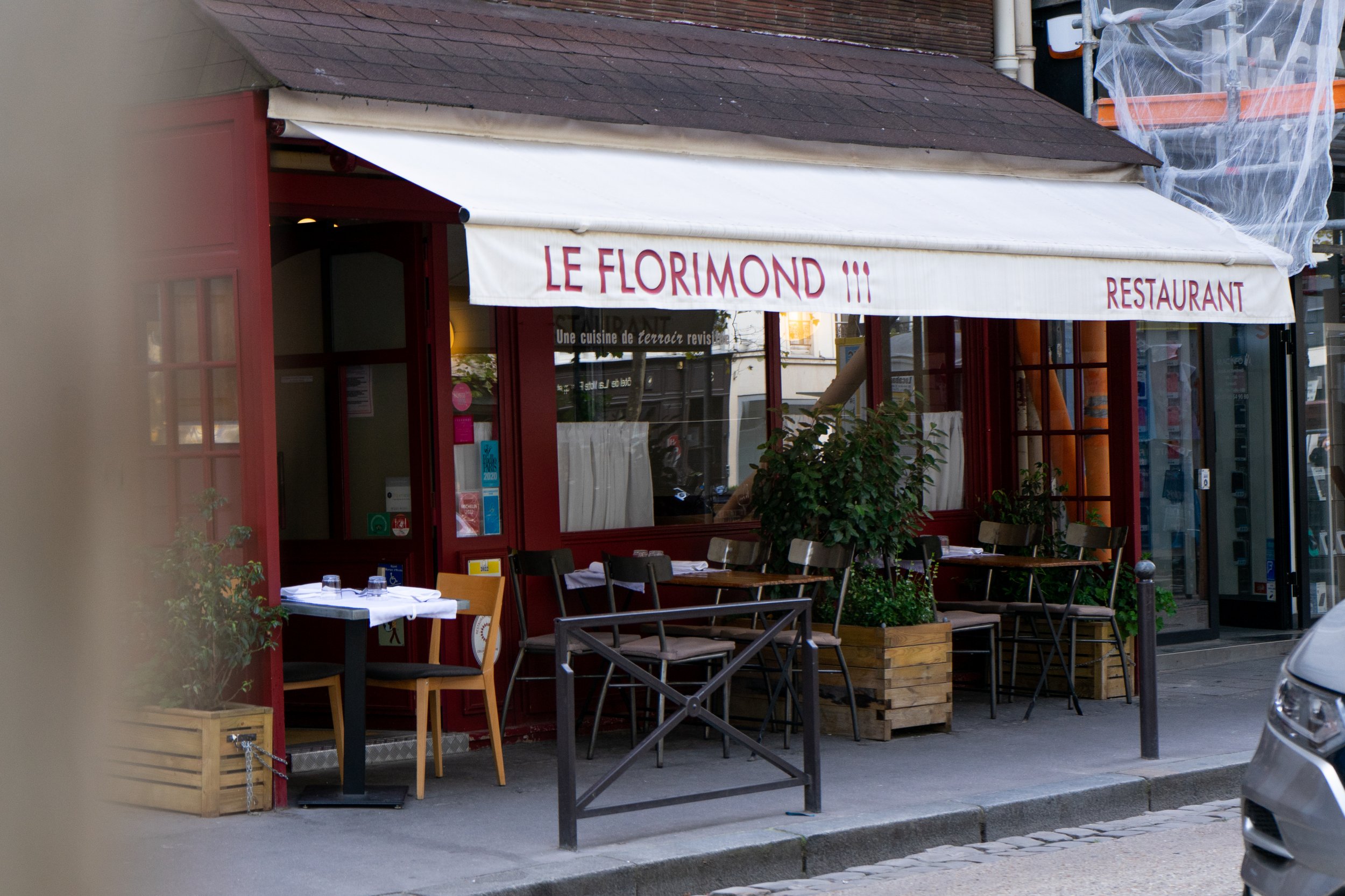 Façade extérieure d'un restaurant nommé 'Le Florimond' avec une terrasse équipée de tables et chaises, décorée de plantes, et une enseigne blanche avec le nom en lettres rouges.