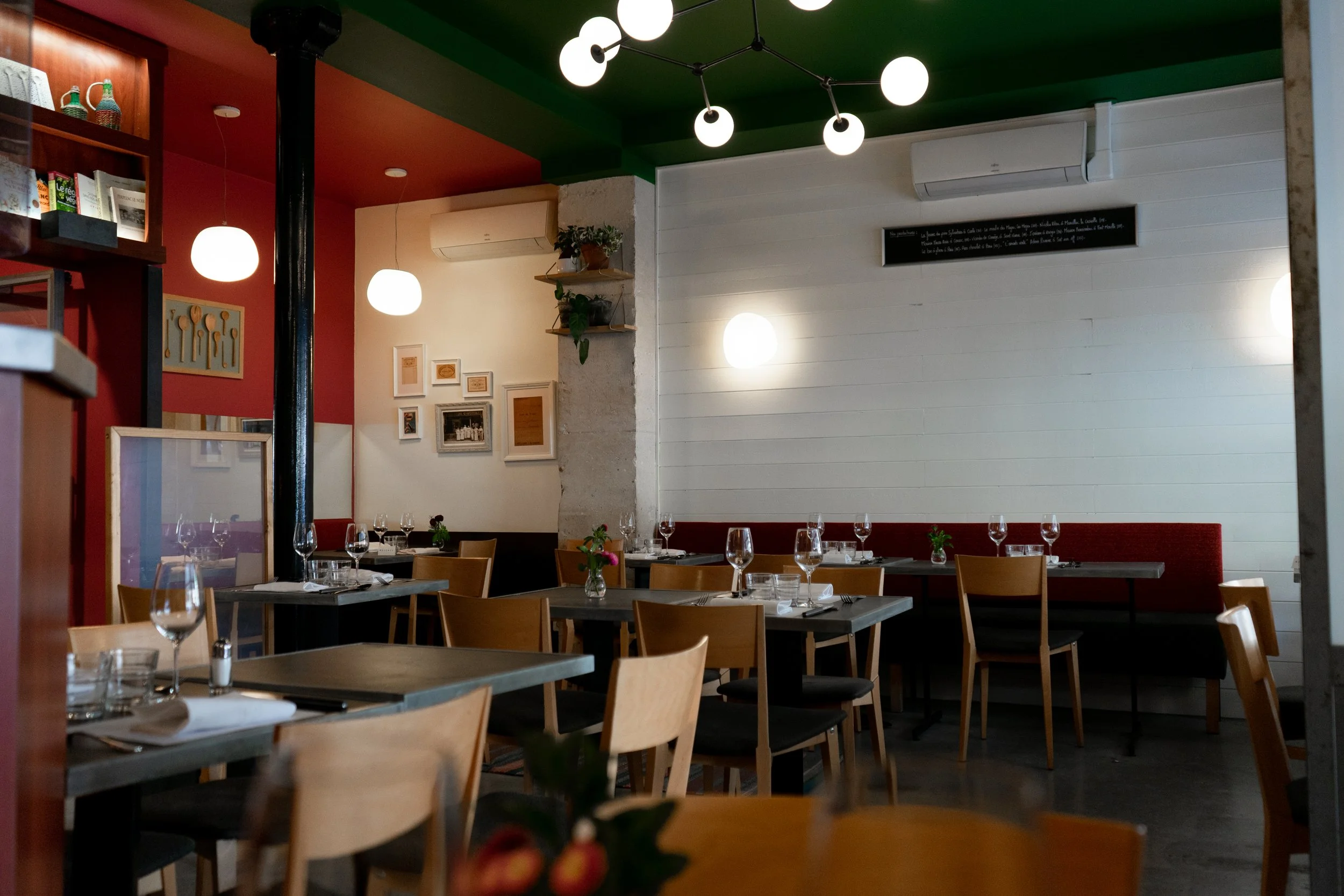 Interior d'un restaurant avec des tables prêtes pour des clients, verres et assiettes, murs décorés avec des tableaux et des plantes en pot, éclairage moderne avec des lampes rondes, plafond vert et murs en blanc et rouge.