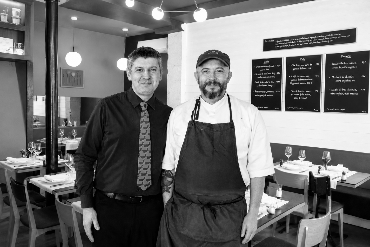 Deux hommes posent dans un restaurant, l'un en chemise noire et la autre en uniforme de chef cuisinier, dans une ambiance moderne avec des tables prêtes pour les clients.