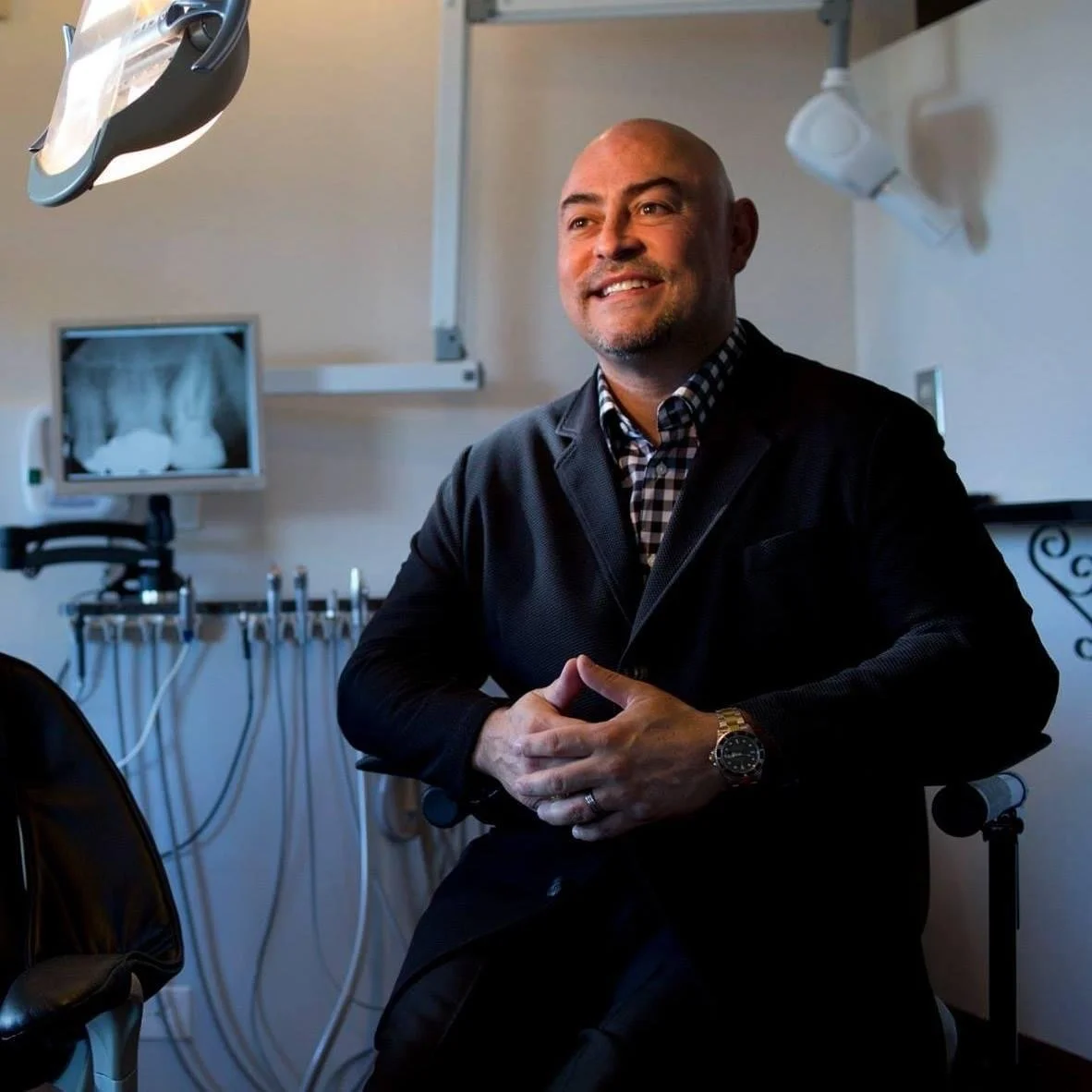 Dr. Chavez in a dental office, smiling, with dental equipment and an X-ray image of teeth on a monitor behind him.