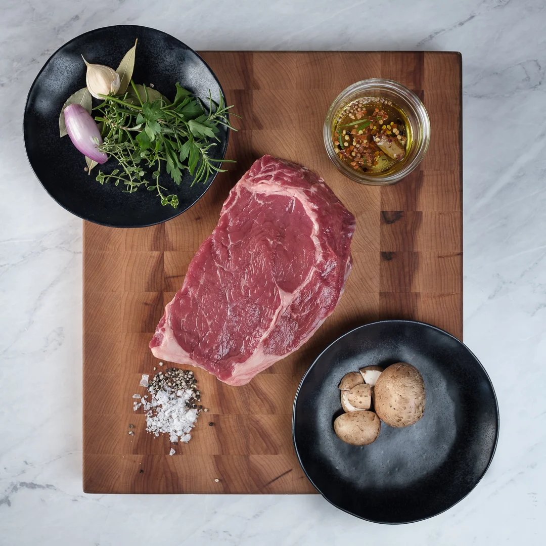 Prime Rib Eye Marinade Prime cut of a beef rib eye on an end grain cutting board featuring marinade ingredients. Marble background.