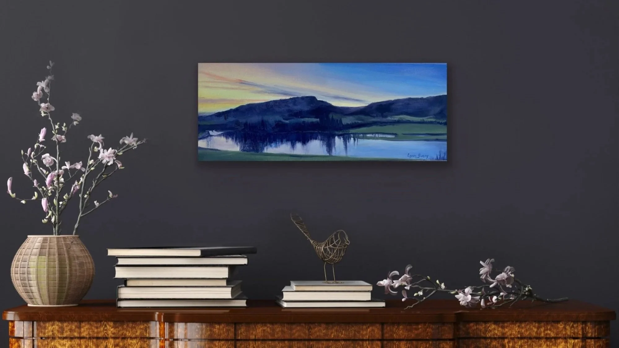 Decorative black wall with landscape painting of a lake and mountains. Wooden table with a vase of pink and white flowers, stacks of black and white books, and a wire sculpture of a bird, with a branch of flowers lying on the table.