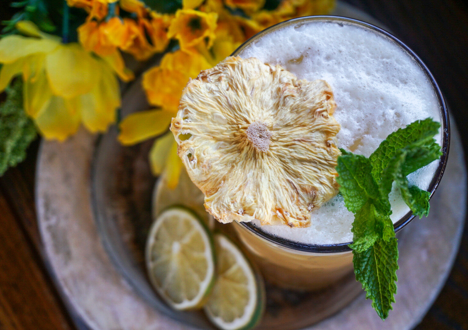  Top view of a frothy decorative cocktail garnished with flowers, citrus slices and a sprig of mint. 