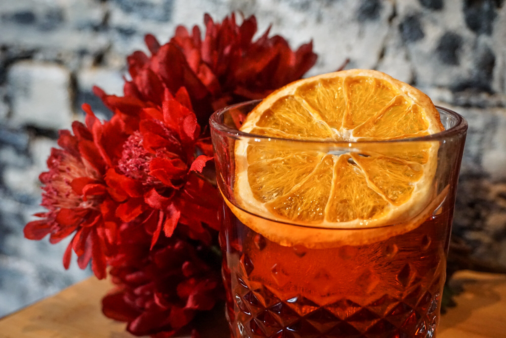  A colorful red cocktail garnished with a dehrydated orange slice next to a red bouquet of flowers. 