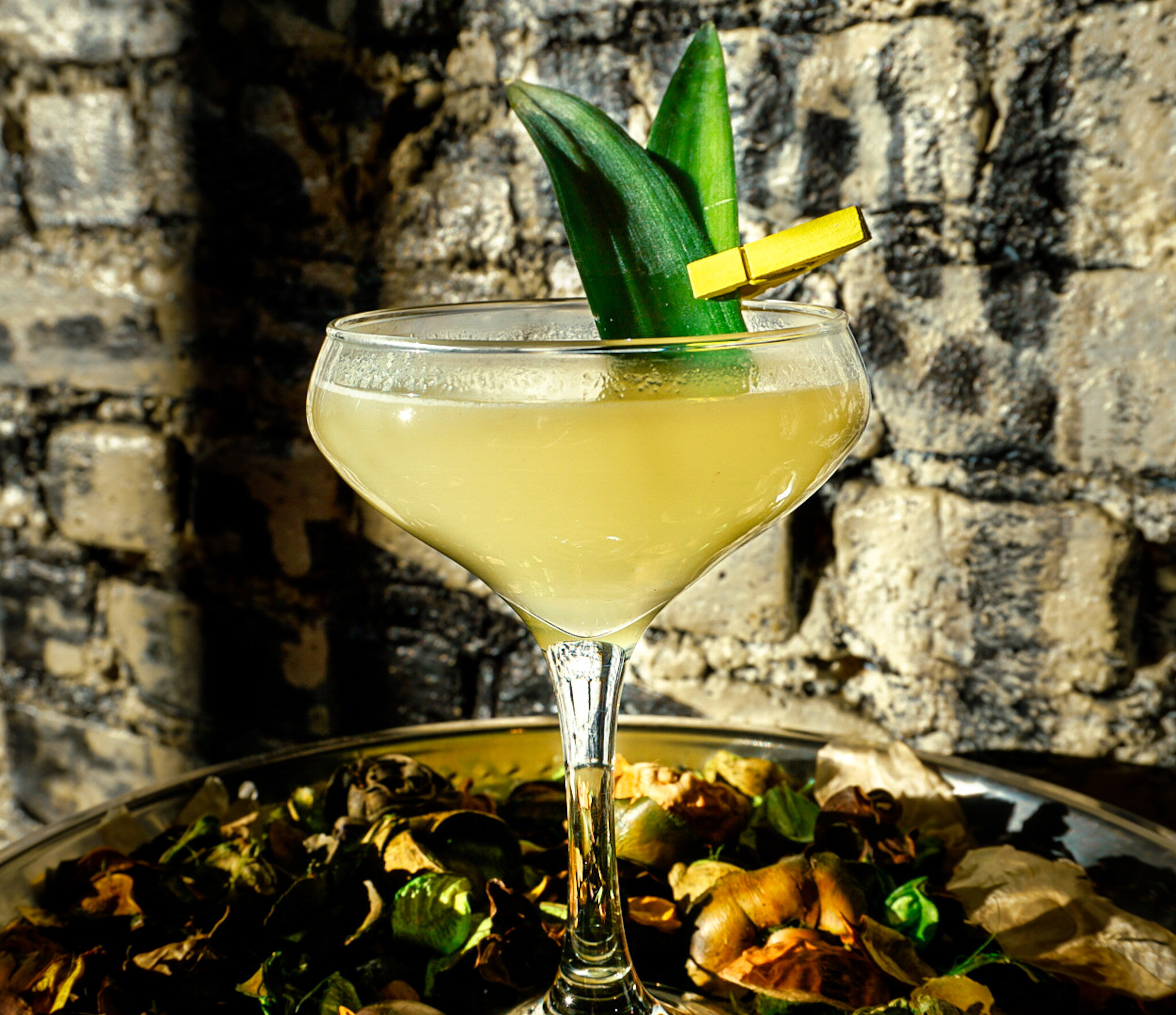  A yellow colored cocktail in a coupe glass garnished with a small, colorful clothes pin holding leaves together. 