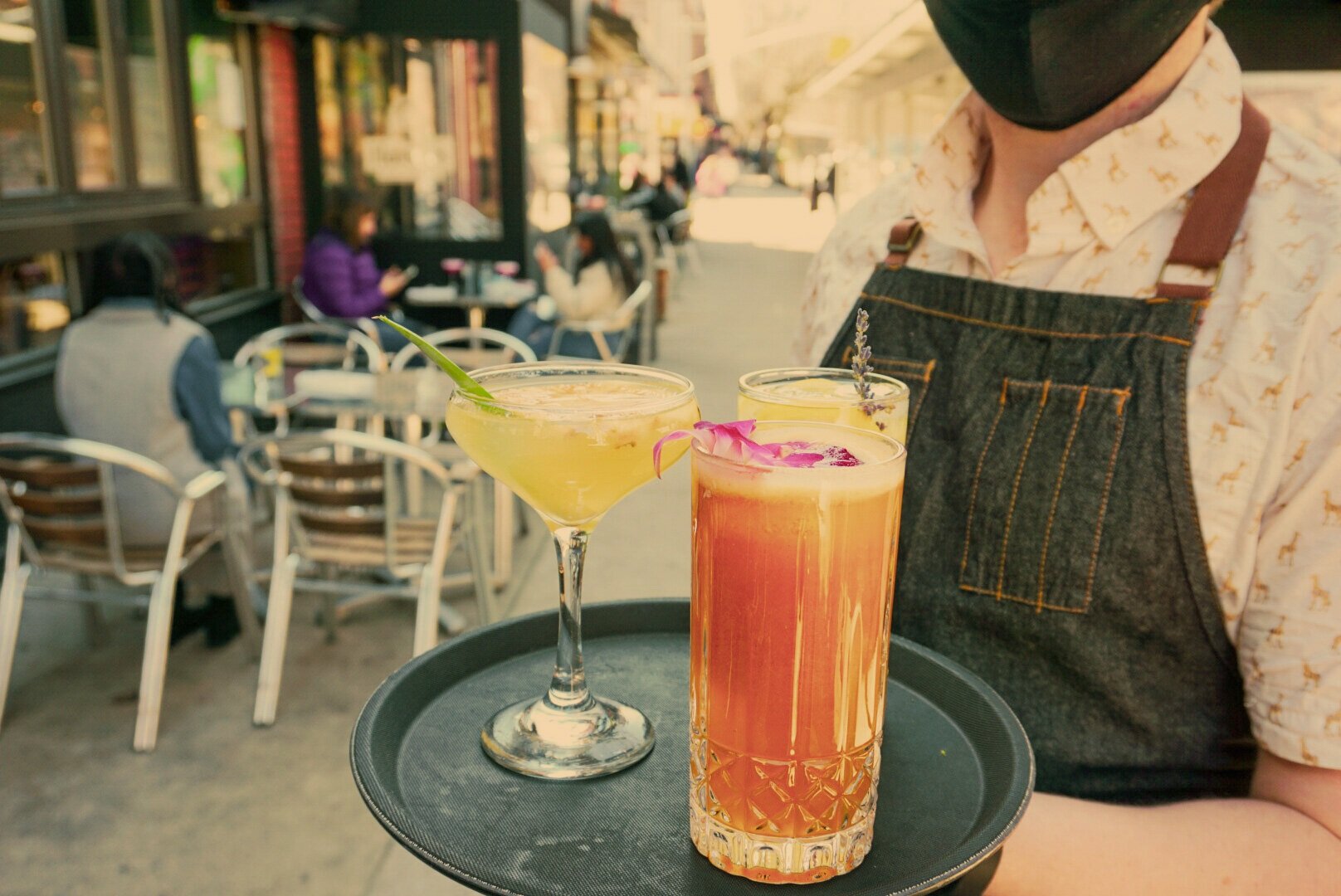  A server holding a tray with three cocktails standing next to outside dining tables. 