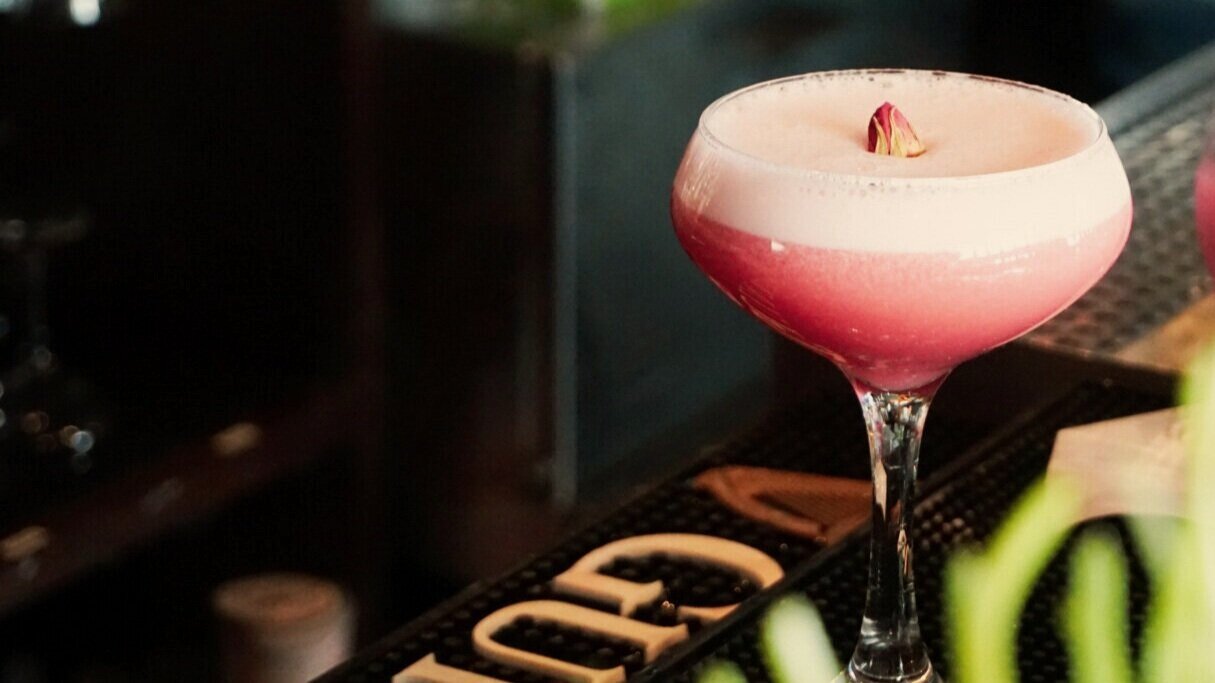  A pink cocktail in a coupe glass sitting on a bar 