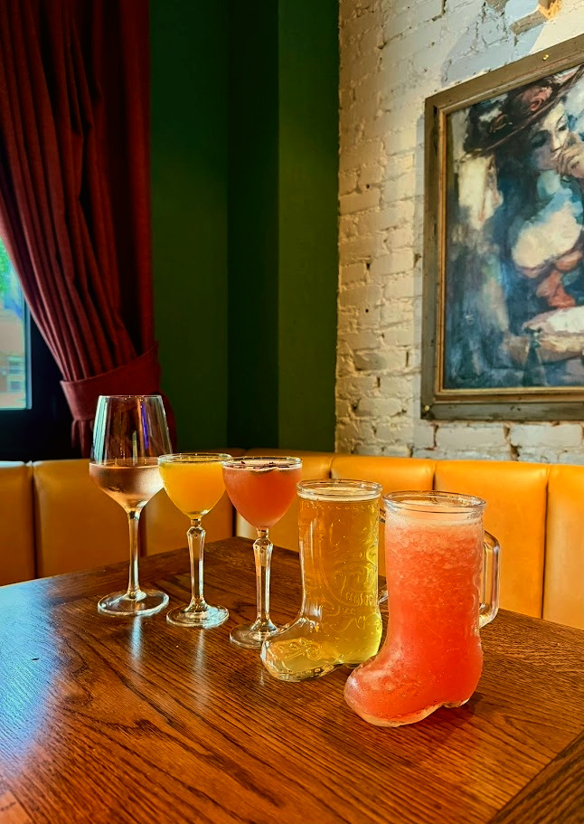 Five glasses, including wine, juices, and beer, and two glass-embossed boots filled with pink-colored drinks on a wooden table in a cozy restaurant with a brick wall, framed artwork, and red curtains.