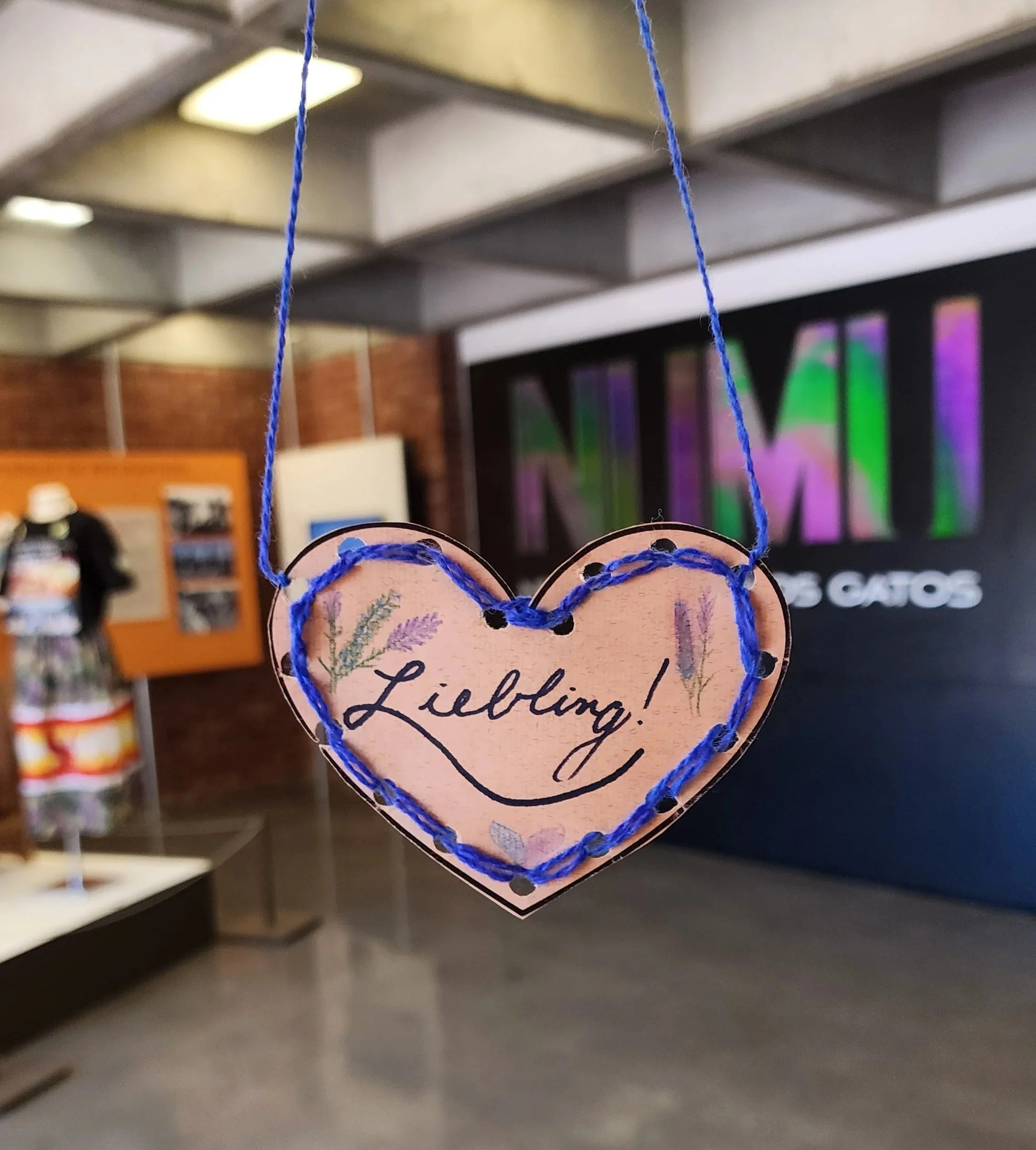 A brown paper heart, embroidered with a blue yarn edge, and attached to a blue yarn necklace. There are illustrations of flowers on the heart and the word "Liebling!