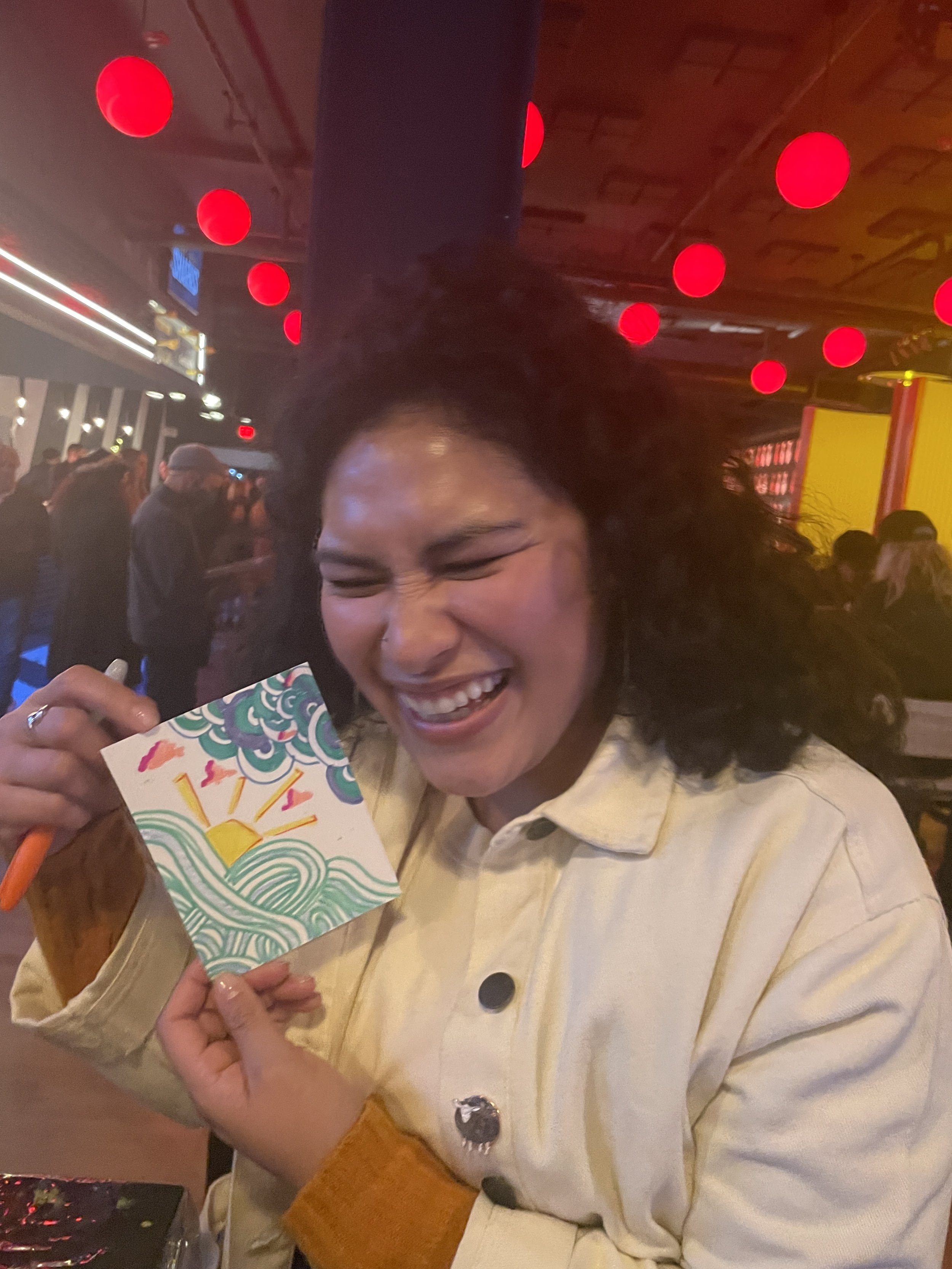 A woman smiling deeply, holding up a postcard she drew with a swirling design