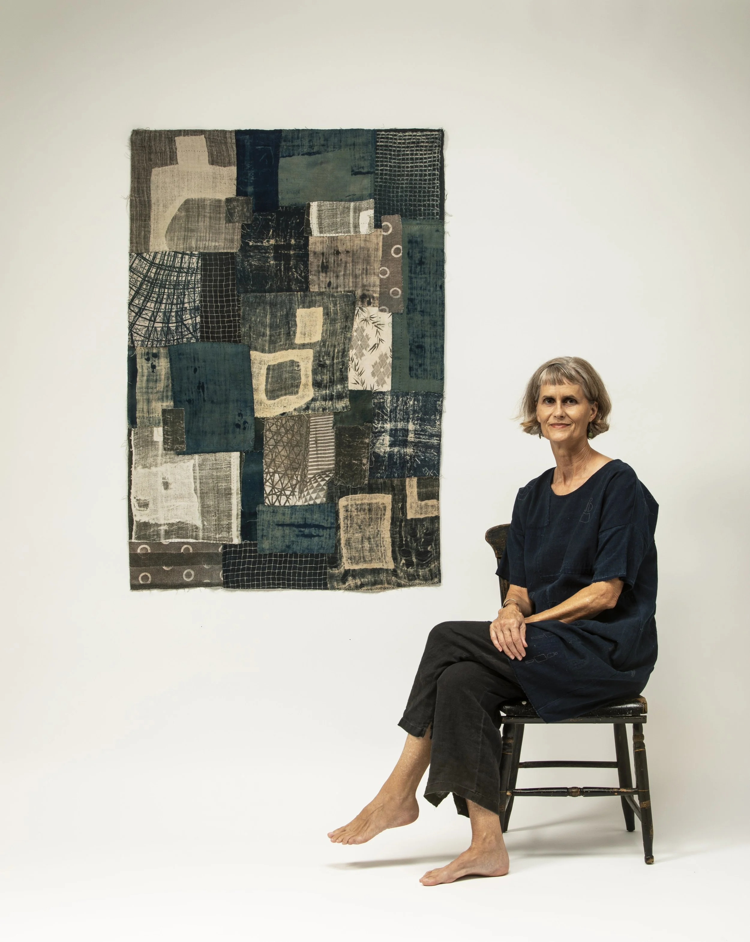 Jody wears a dark colored outfit and sits on a wooden chair in front of a blue and tan quilt hung on the wall