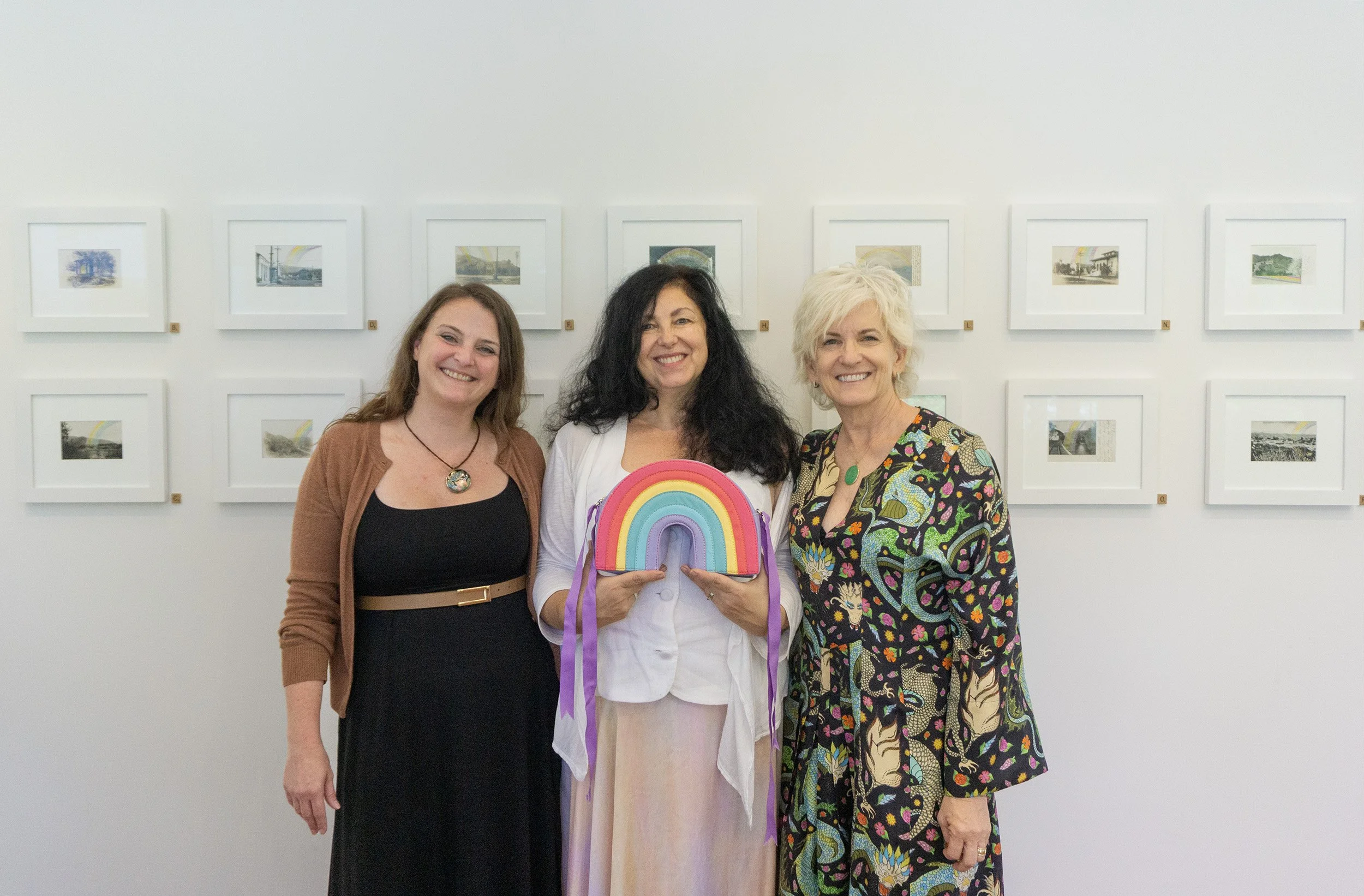 Three women pose in front of framed artworks. Marie, in the middle, holds up a purse made to look like a rainbow