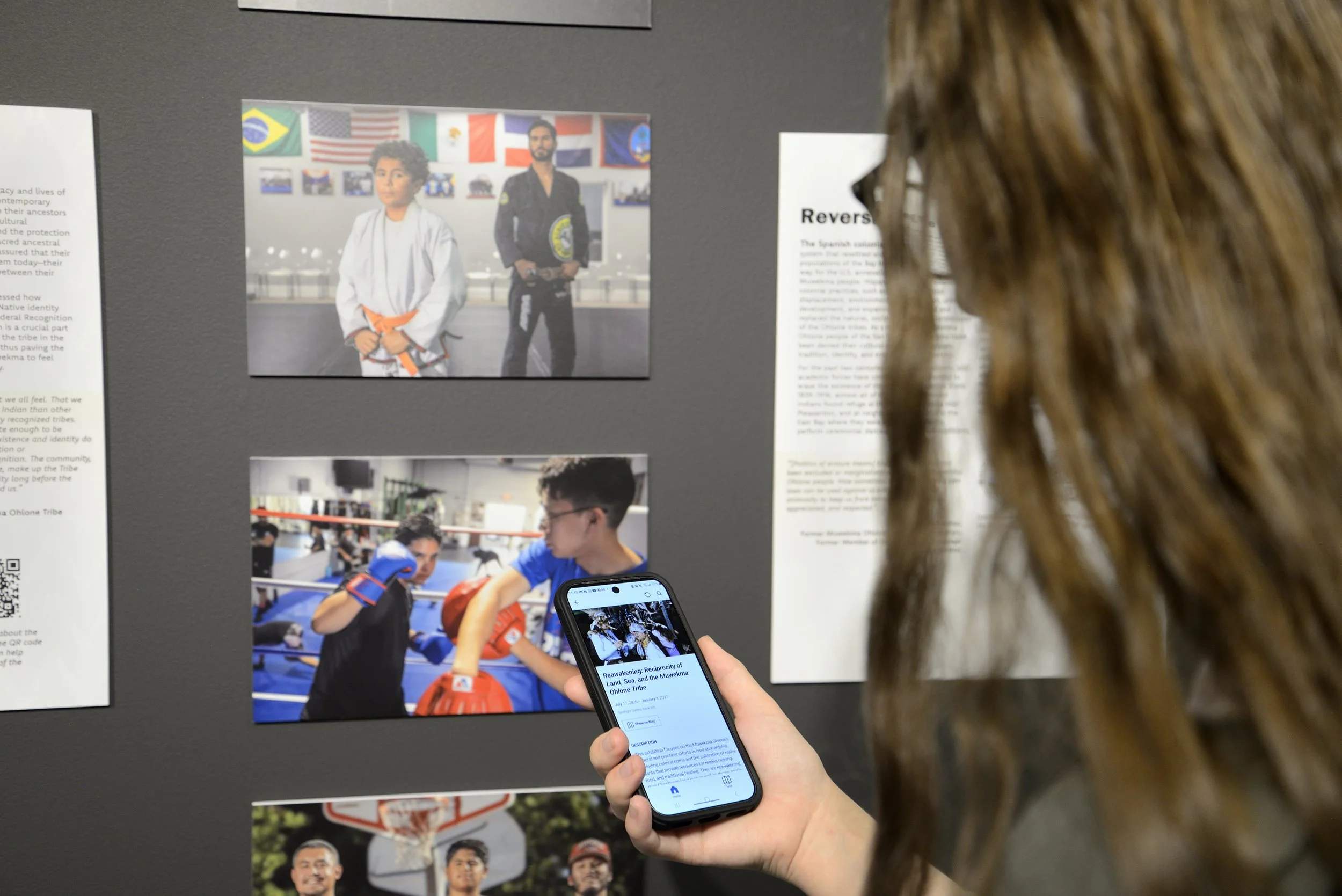 A person with long brown hair holds a smartphone displaying text and an image, looking at a museum exhibit. On the dark gray wall, several photographs show young men in martial arts uniforms, boxing, and playing basketball, alongside text panels.