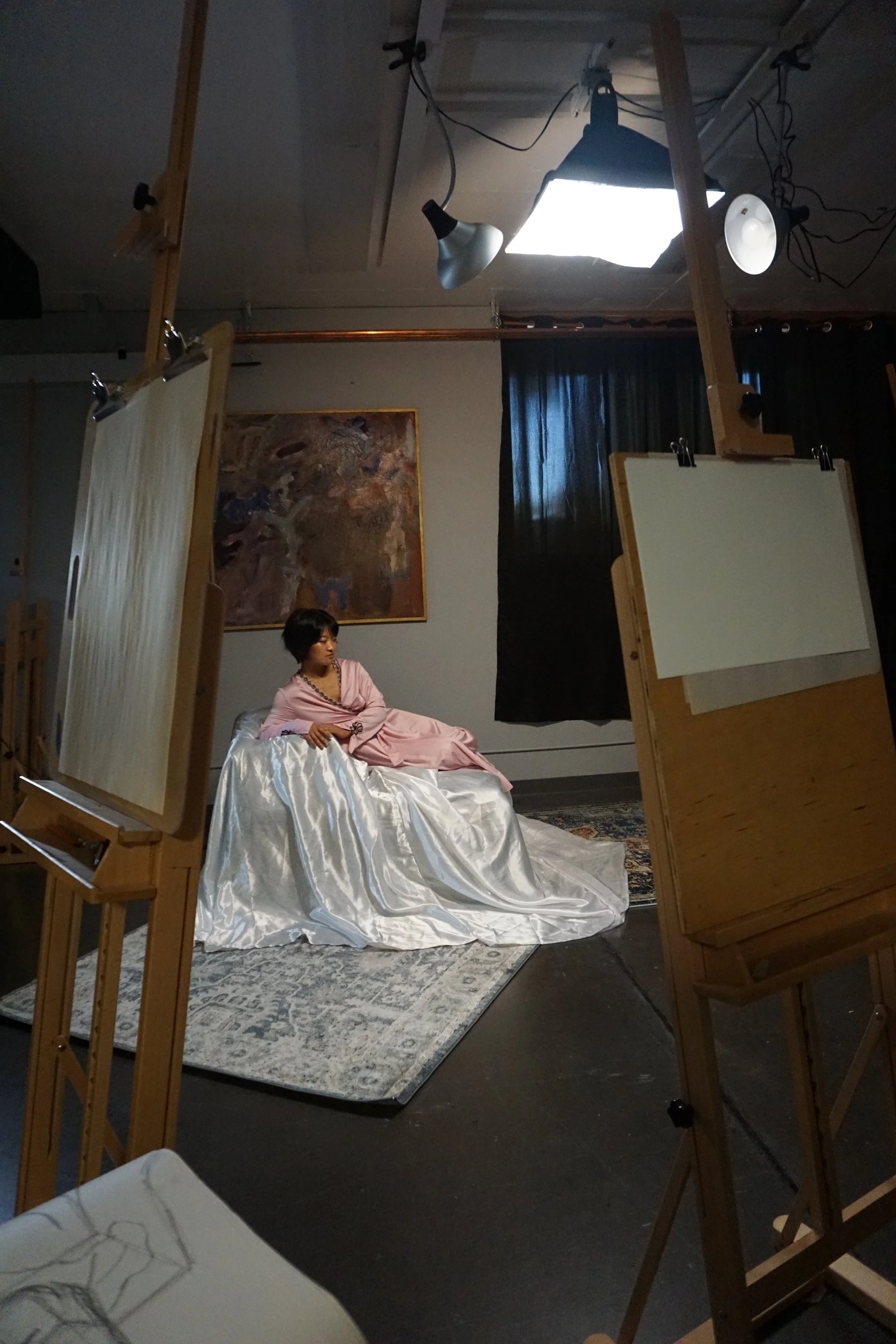 A model in a pink robe, laying on a satin structure, surrounded by easels