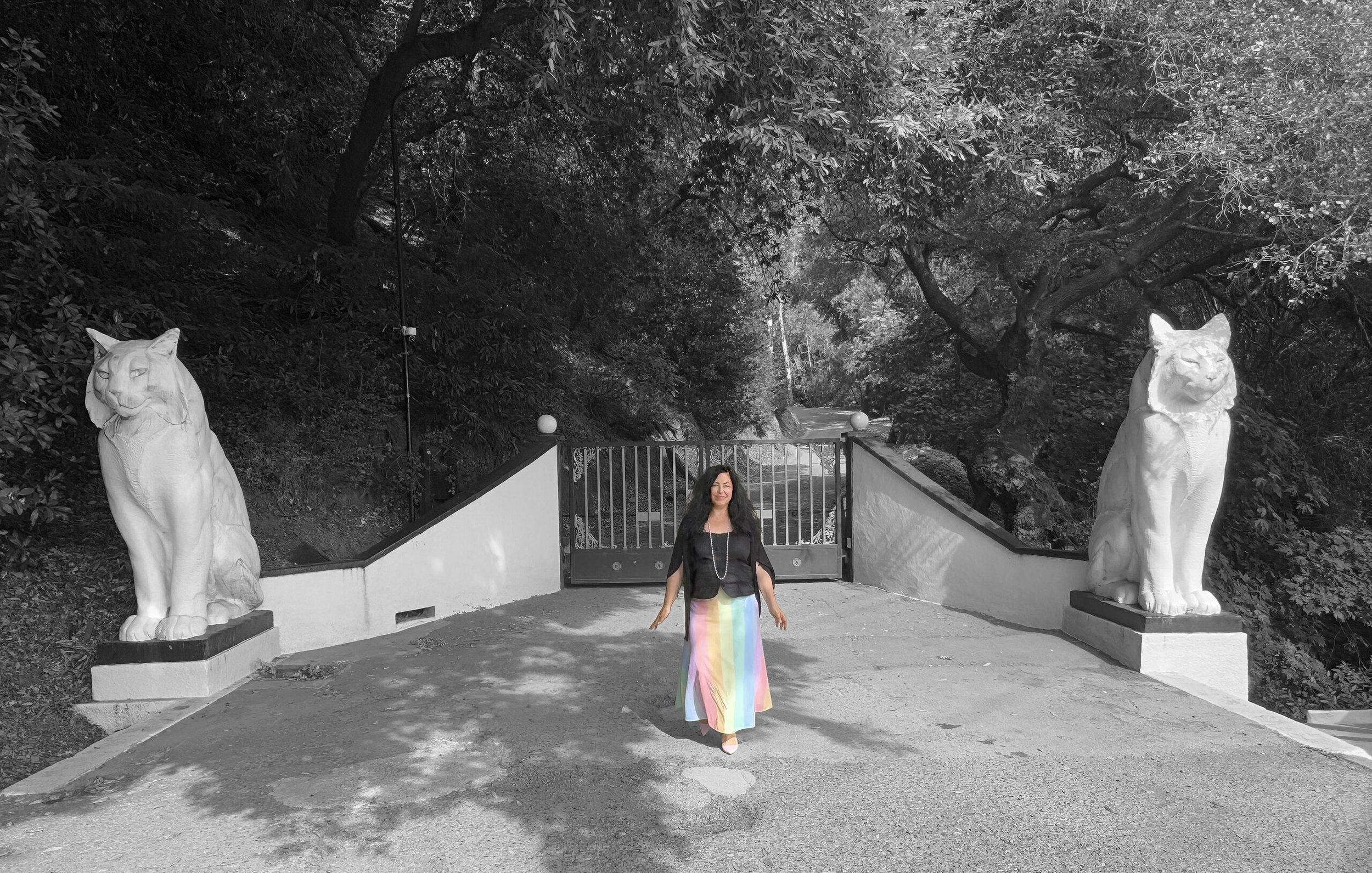 Marie stands in front of "The Cats". She wears a rainbow skirt. The photo has been edited to be black and white other than her.