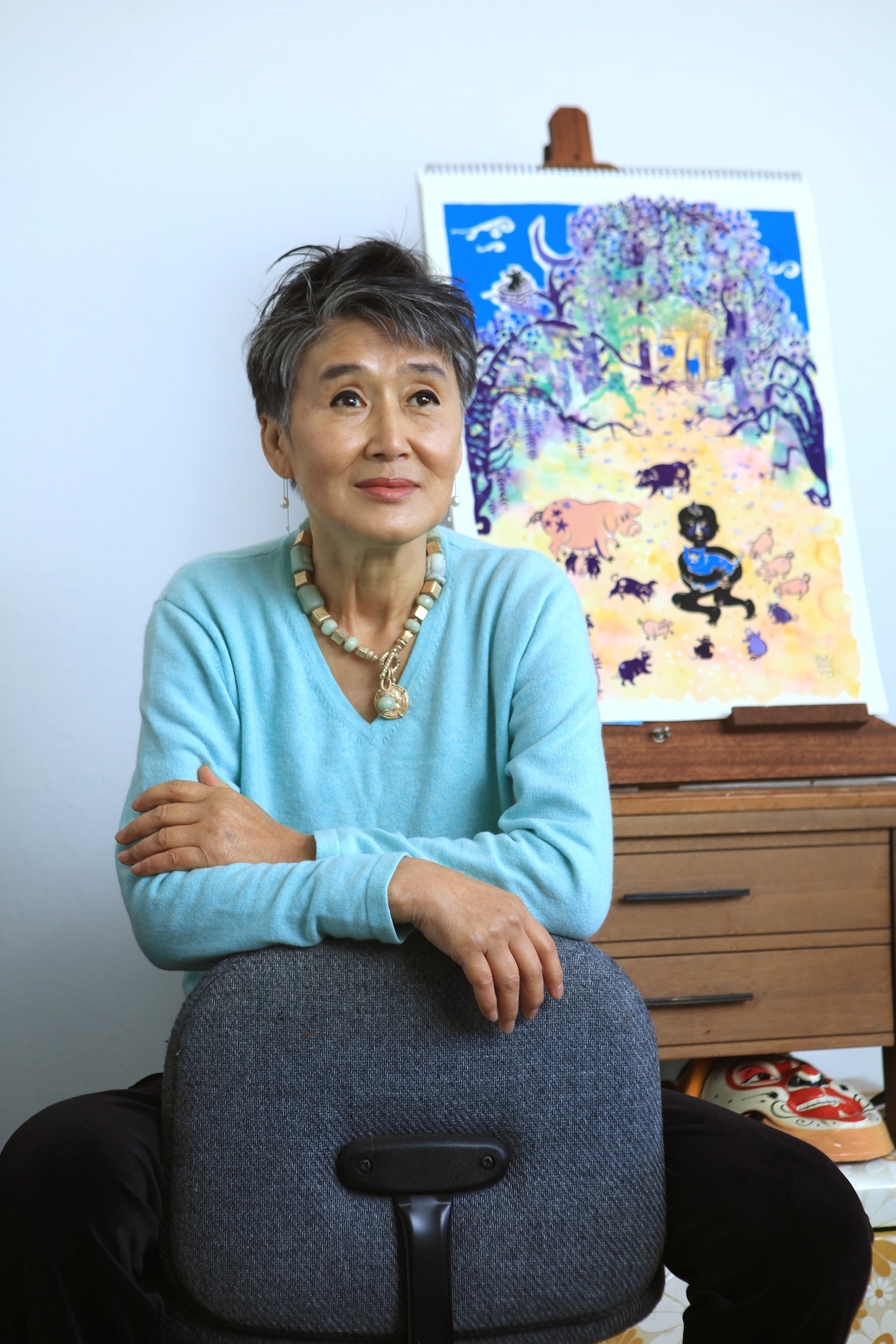 Belle, a woman with cropped black hair, sits backwards on an office hair. She wears a blue shirt and beaded necklace. Behind her sits a set of drawers, topped with a painting of a forest scene, and a mask sits underneath
