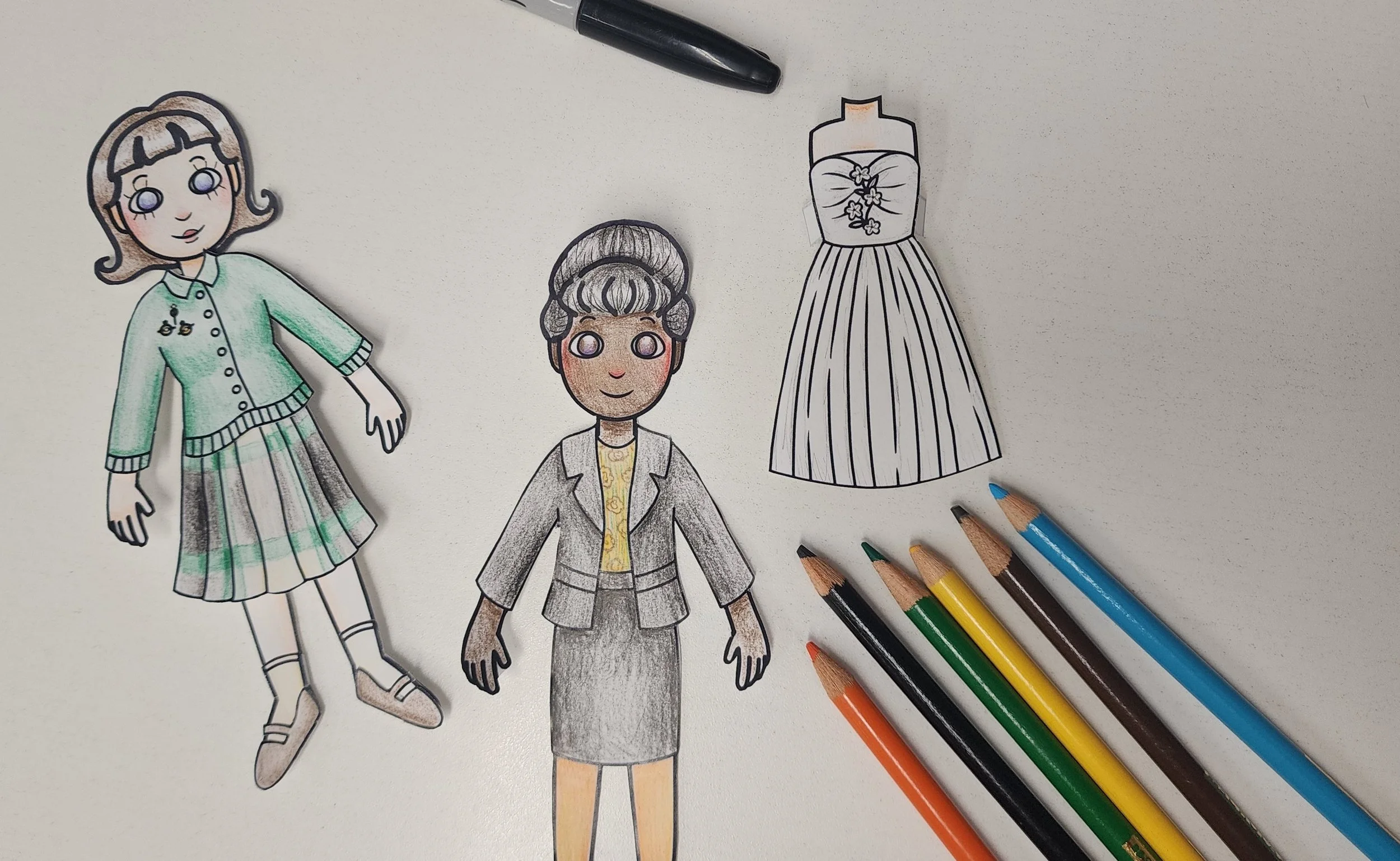 Paper dolls in 60s clothing, markers, and colored pencils on a white table