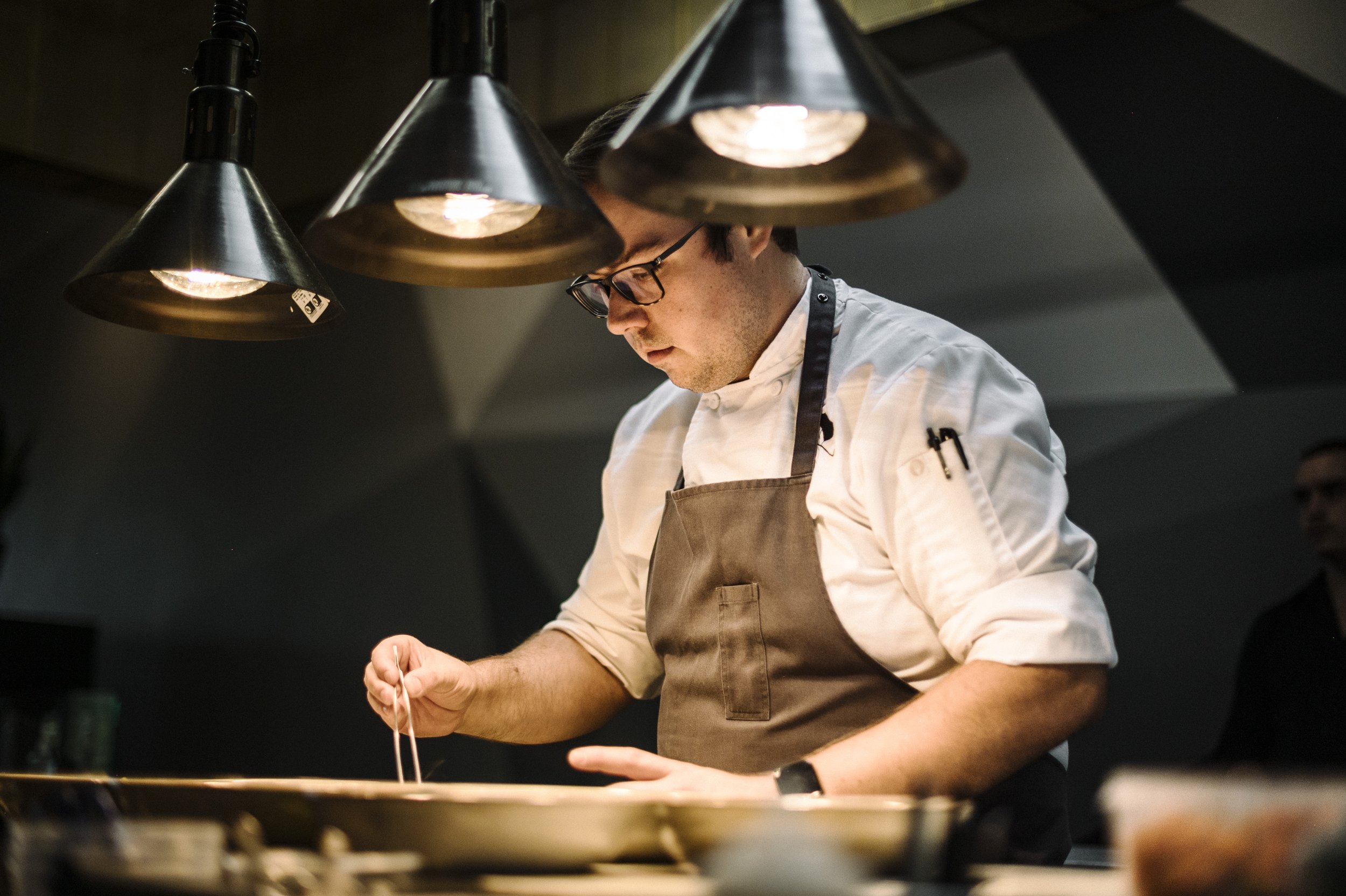 Chef wearing glasses and a white uniform, working in a professional kitchen, illuminated by overhead pendant lights.