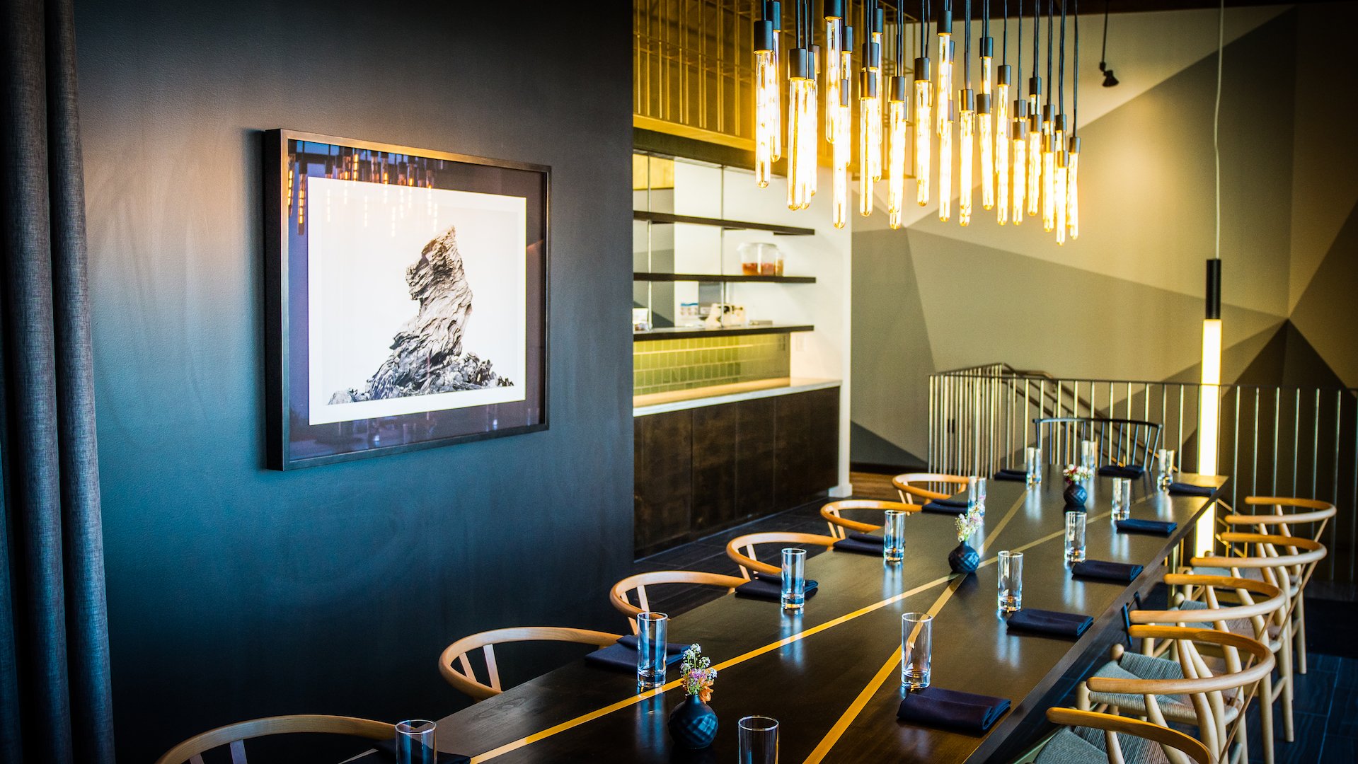 Modern dining room with a long dark table set with glasses, black napkins, and small flower arrangements, surrounded by wooden chairs, with stylish hanging lights above and artwork on the wall.