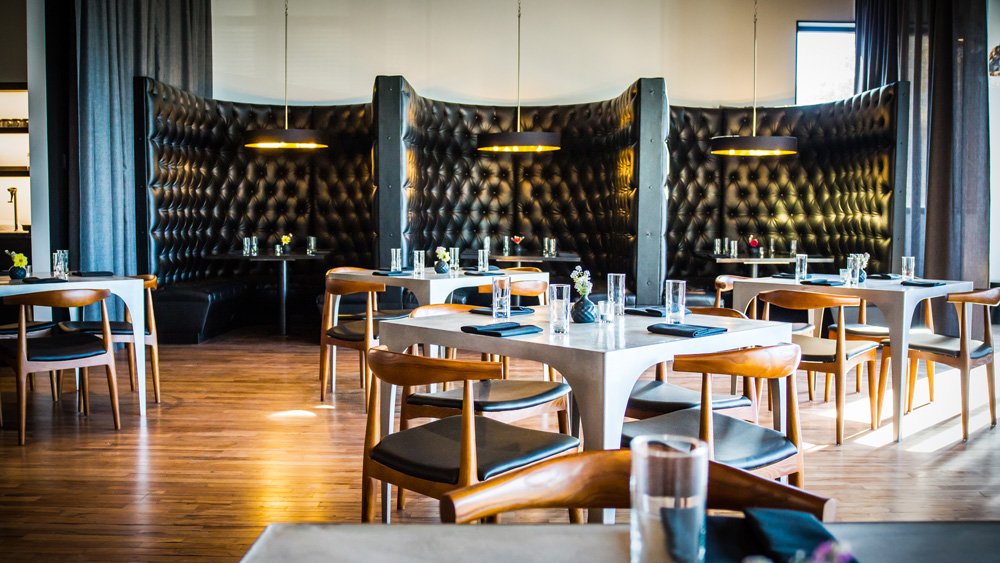 Empty restaurant with modern decor, dark leather booths, white tables, wooden chairs, and pendant lighting.