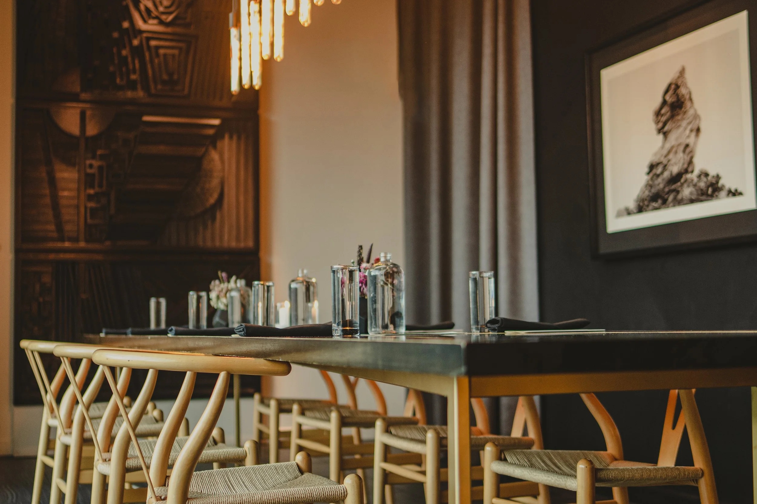 A modern dining room with a long table set with glassware, black napkins, and small flower arrangements. Wooden chairs are placed around the
