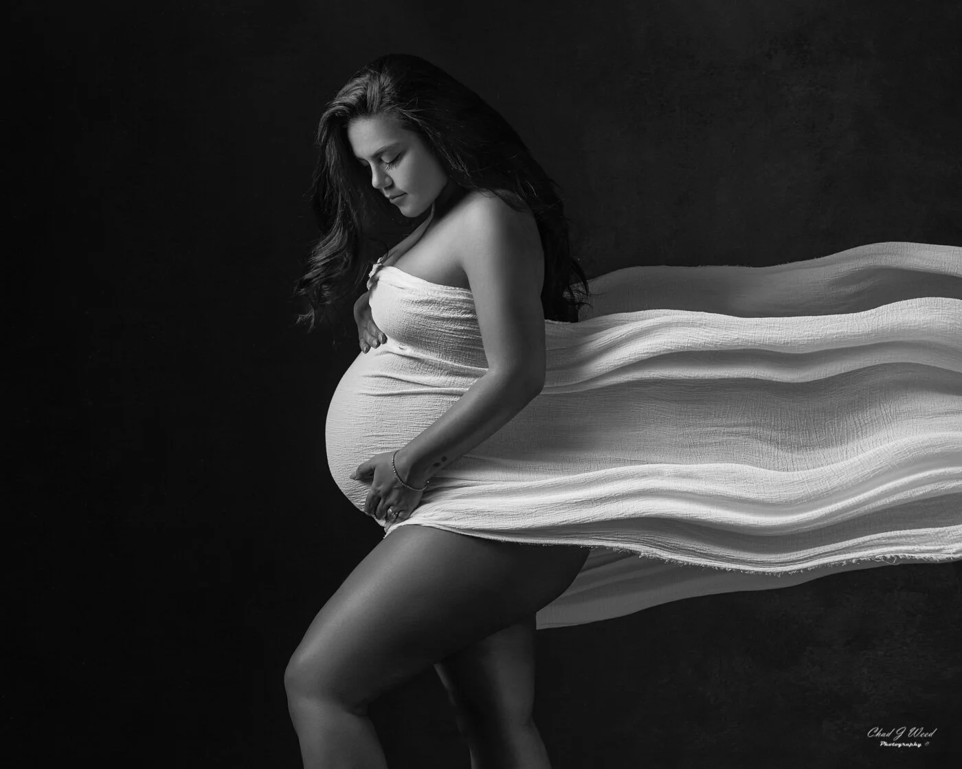 Pregnant woman wrapped in flowing material looking down at her belly during a maternity photo session in Gilbert