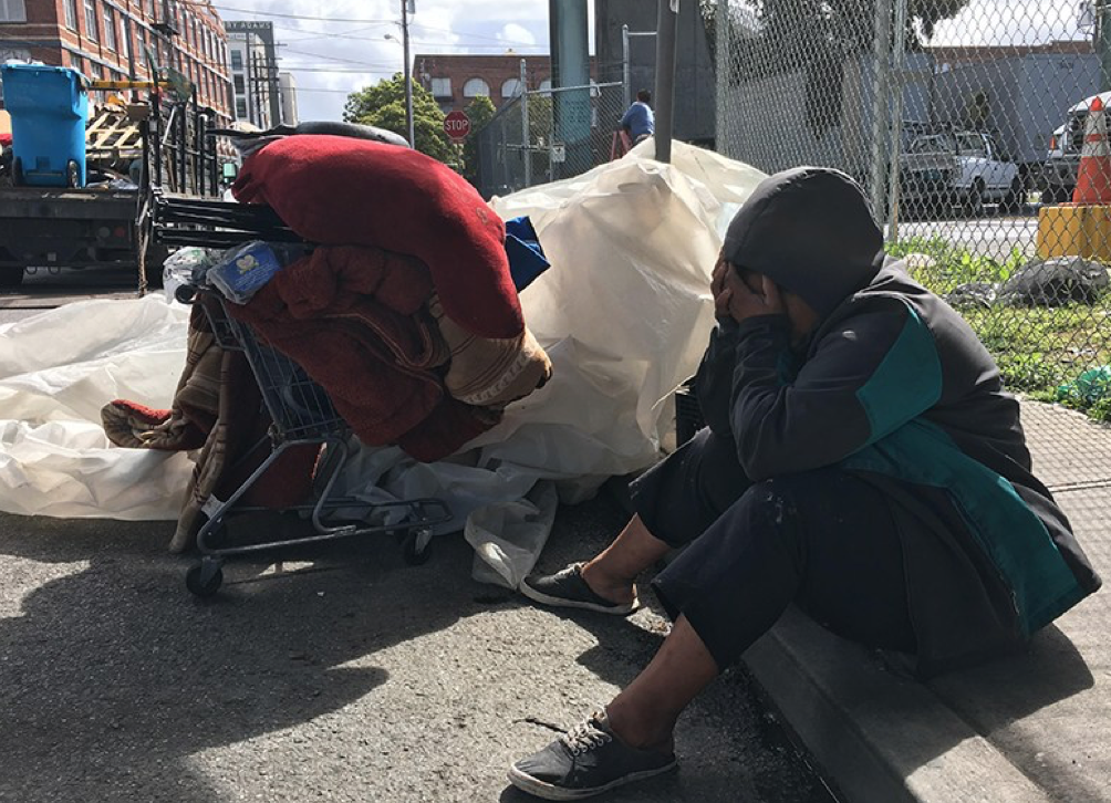 Shy, who lives in a homeless encampment on Alameda Street at San Bruno Avenue in San Francisco, sits on the curb while a Public Works cleaning crews sorts through her belongings. Photo by Laura Waxmann.