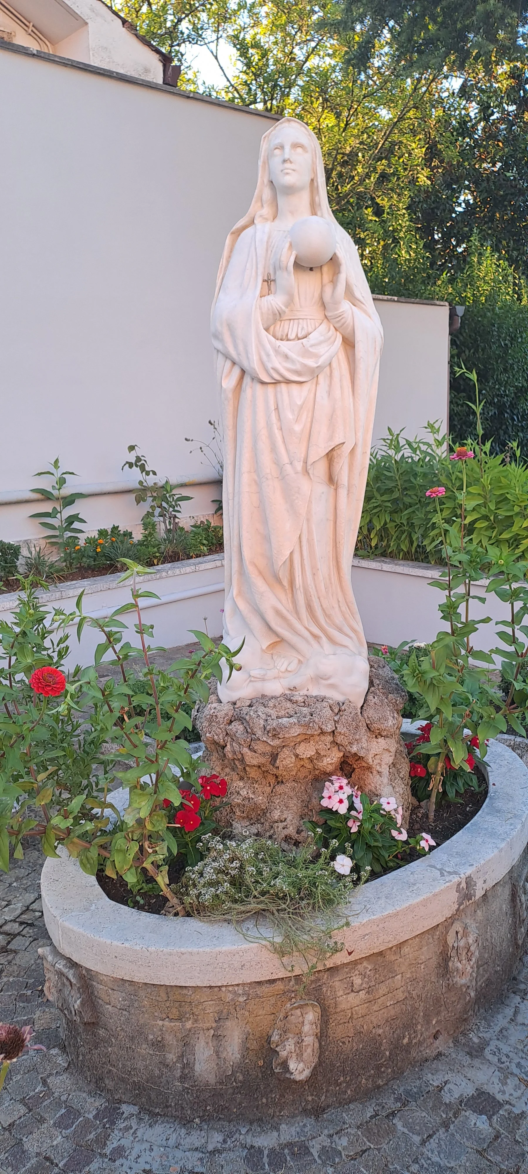 A white marble statue of a woman holding a globe, standing on a stone base surrounded by flowering plants in a circular flower bed.