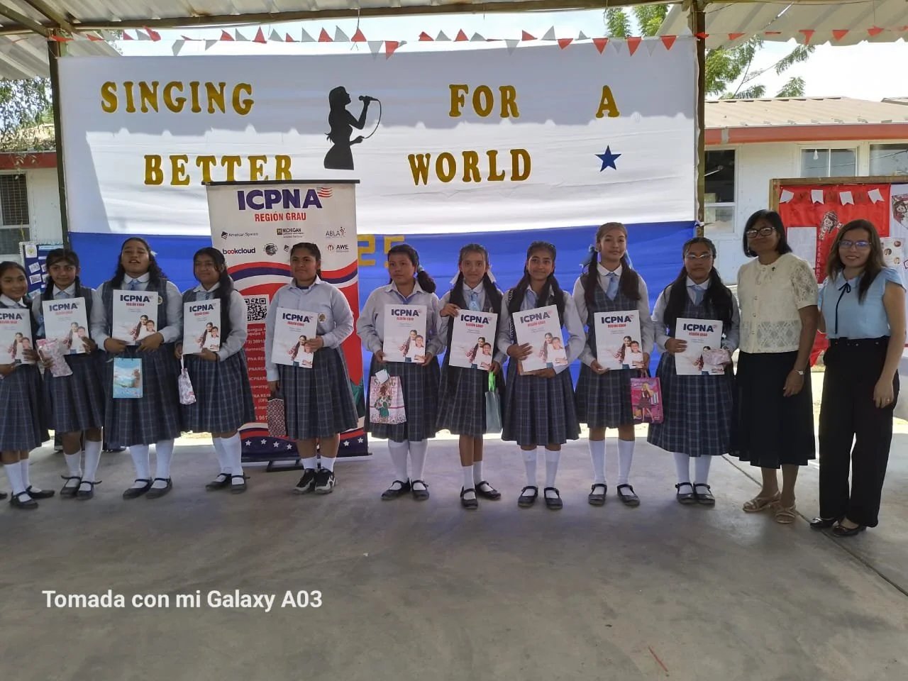 Group of students and teachers standing in front of a banner that reads 'Singing for a better world,' holding pamphlets or certificates, during an event in an outdoor covered area.
