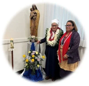 Two women standing beside a pillar with a religious statue, flowers, and an Hawaiian lei, indoors with white walls and curtains.