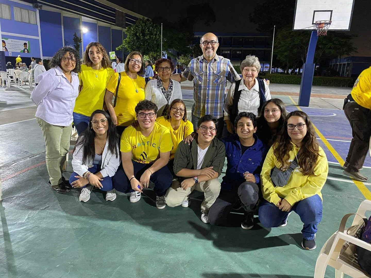 Group of 13 smiling people, some wearing yellow shirts, gathered on an outdoor basketball court at night, with a few chairs and tables in the background.