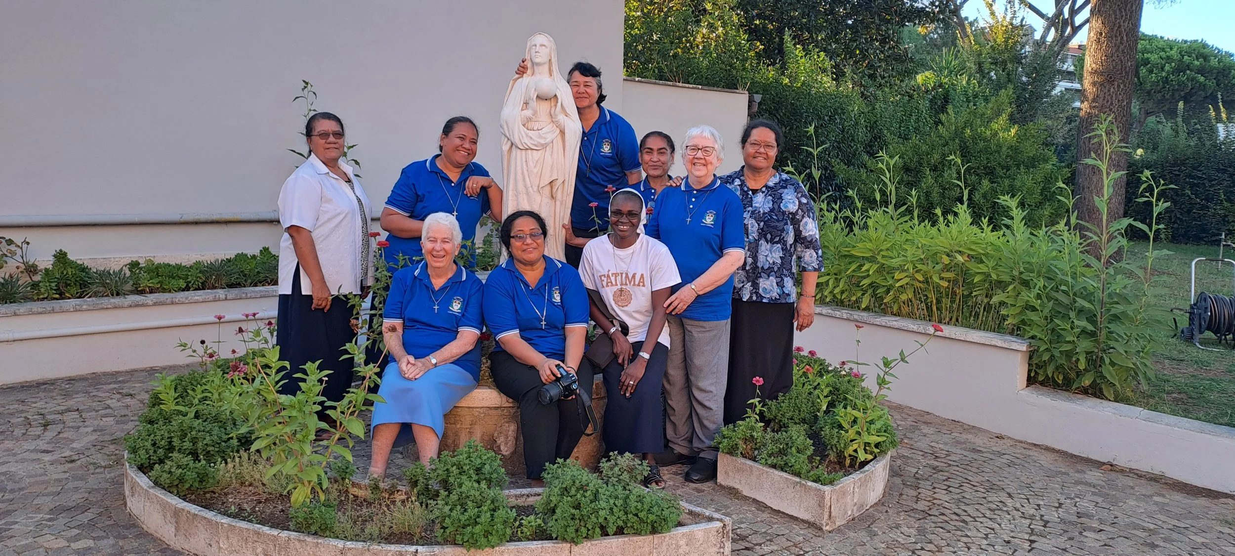 Group of ten women, some wearing blue shirts with a crest and others in casual clothing, standing and sitting around a stone statue of Jesus holding a heart, in an outdoor garden with greenery and flowers.