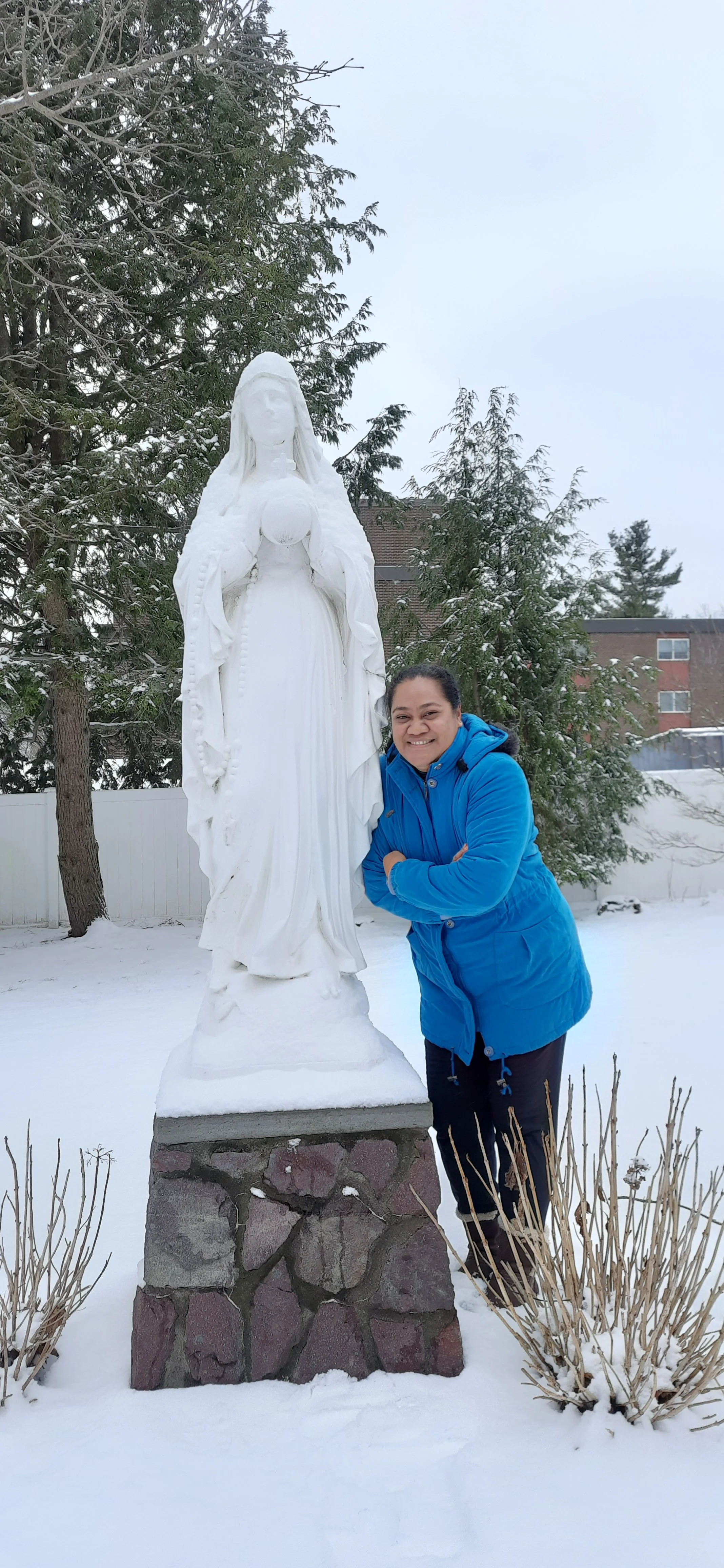 A woman in a bright blue winter jacket smiles and poses next to a white statue of the Virgin Mary outdoors in a snowy yard, with trees and a building in the background.
