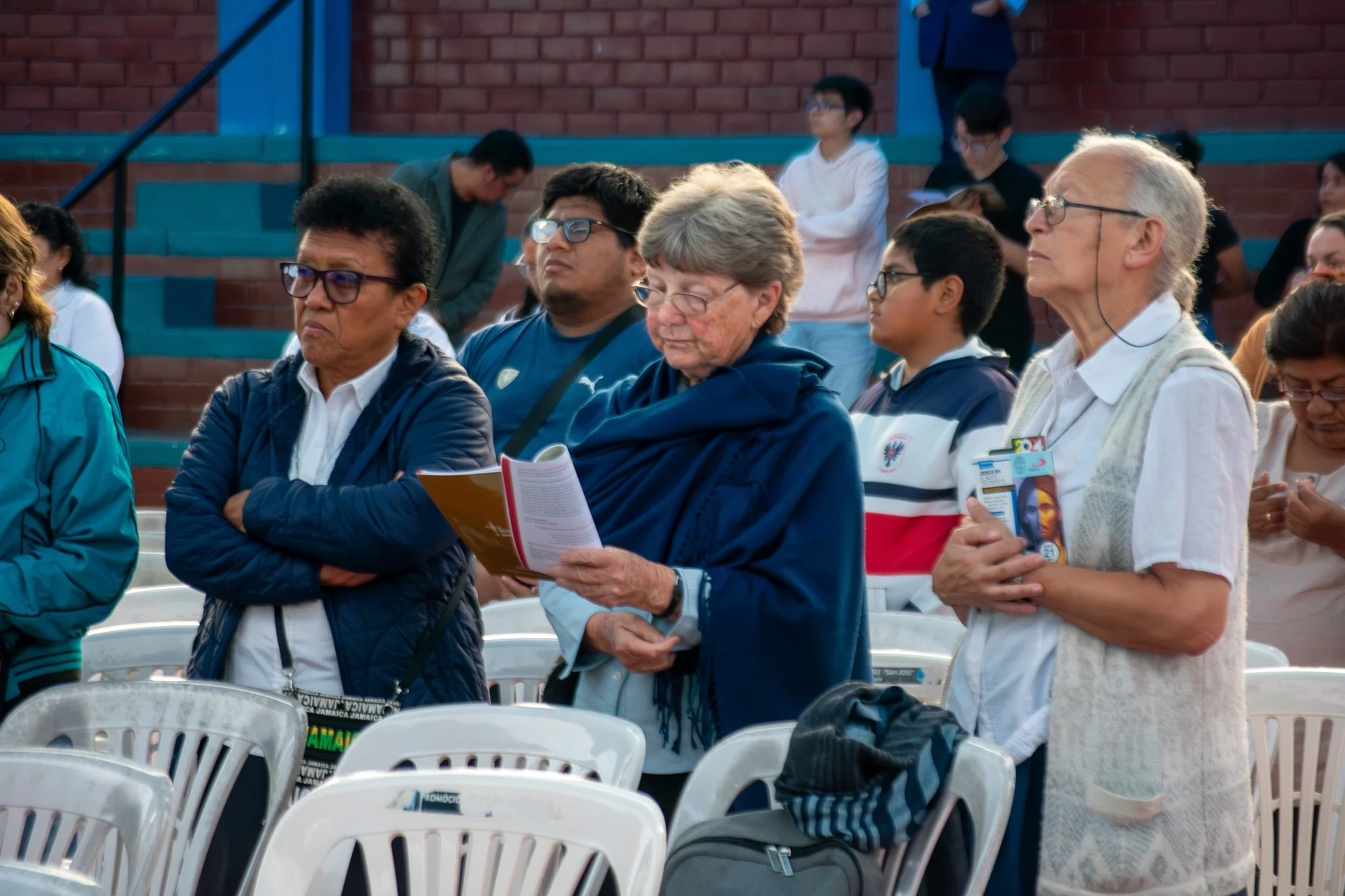 A group of people standing with eyes closed and reading materials, likely participating in a religious or spiritual gathering, with some holding prayer cards or booklets, in an outdoor setting with chairs and steps in the background.