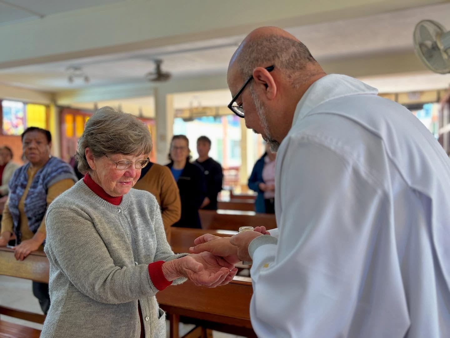 A priest is giving communion to an elderly woman during a religious service in a church, with several people in the background.