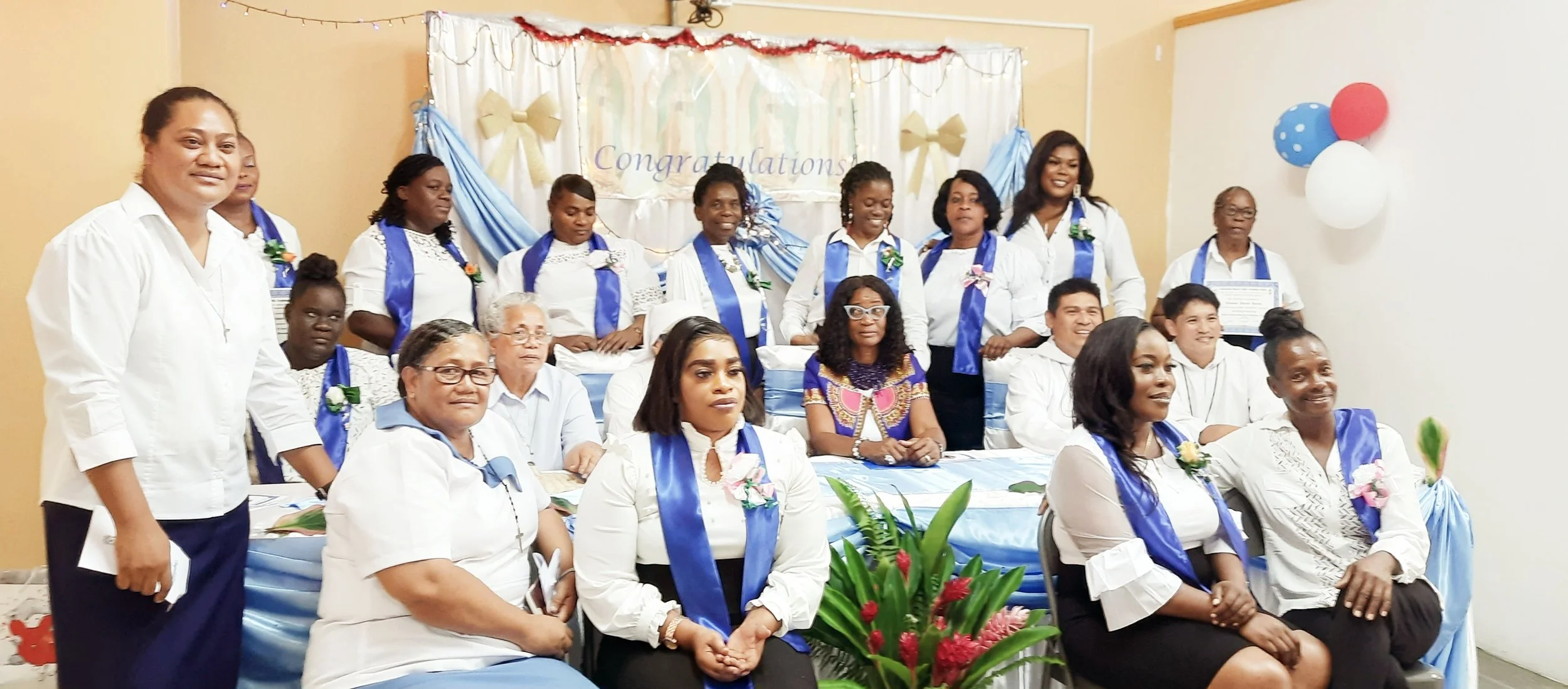 Group of people at a celebration or graduation event, dressed in white and blue attire, with some wearing sashes and corsages, in front of a decorated backdrop with the word 'Congratulations.'
