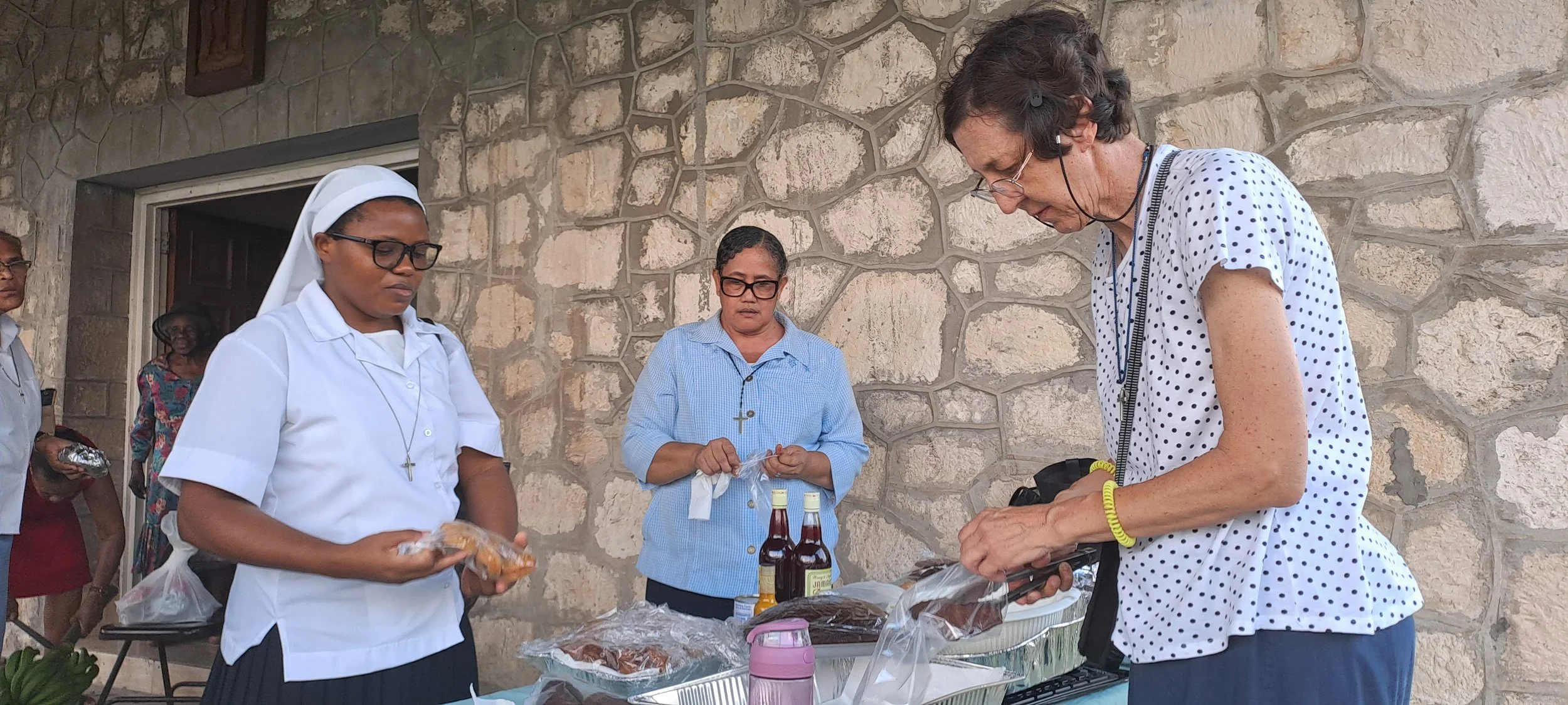 A group of women, including a nun, standing outside at a table with food and bottles, engaging in a food distribution event.