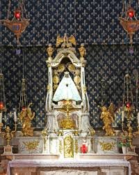 An ornate religious altar with gold accents, featuring a central statue cloaked in white, flanked by smaller statues and candles.