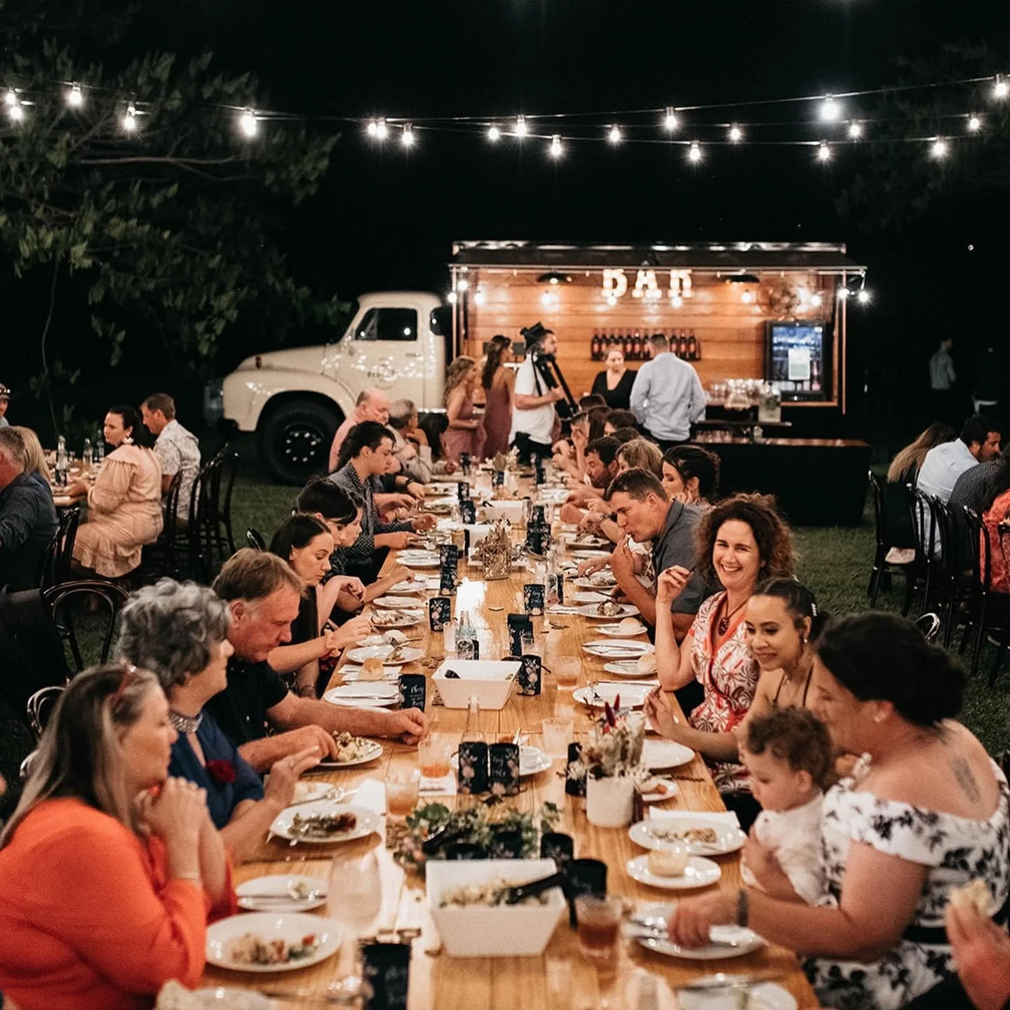 Guests dining at a long outdoor table at night with a food truck and bar in the background, illuminated by string lights.