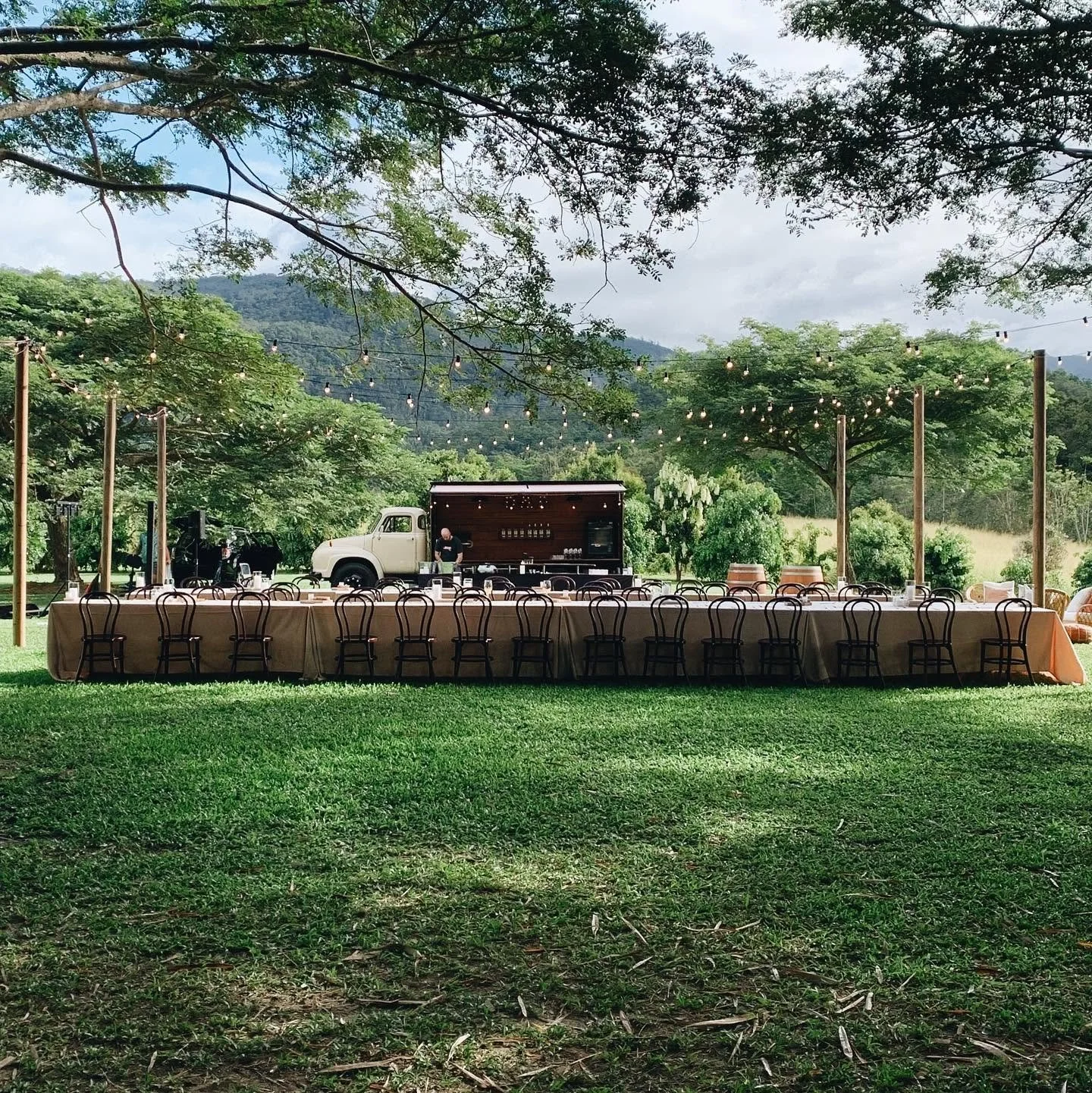 Long outdoor banquet table set up with chairs under a large tree with string lights, with a vintage truck and lush green mountains in the background.