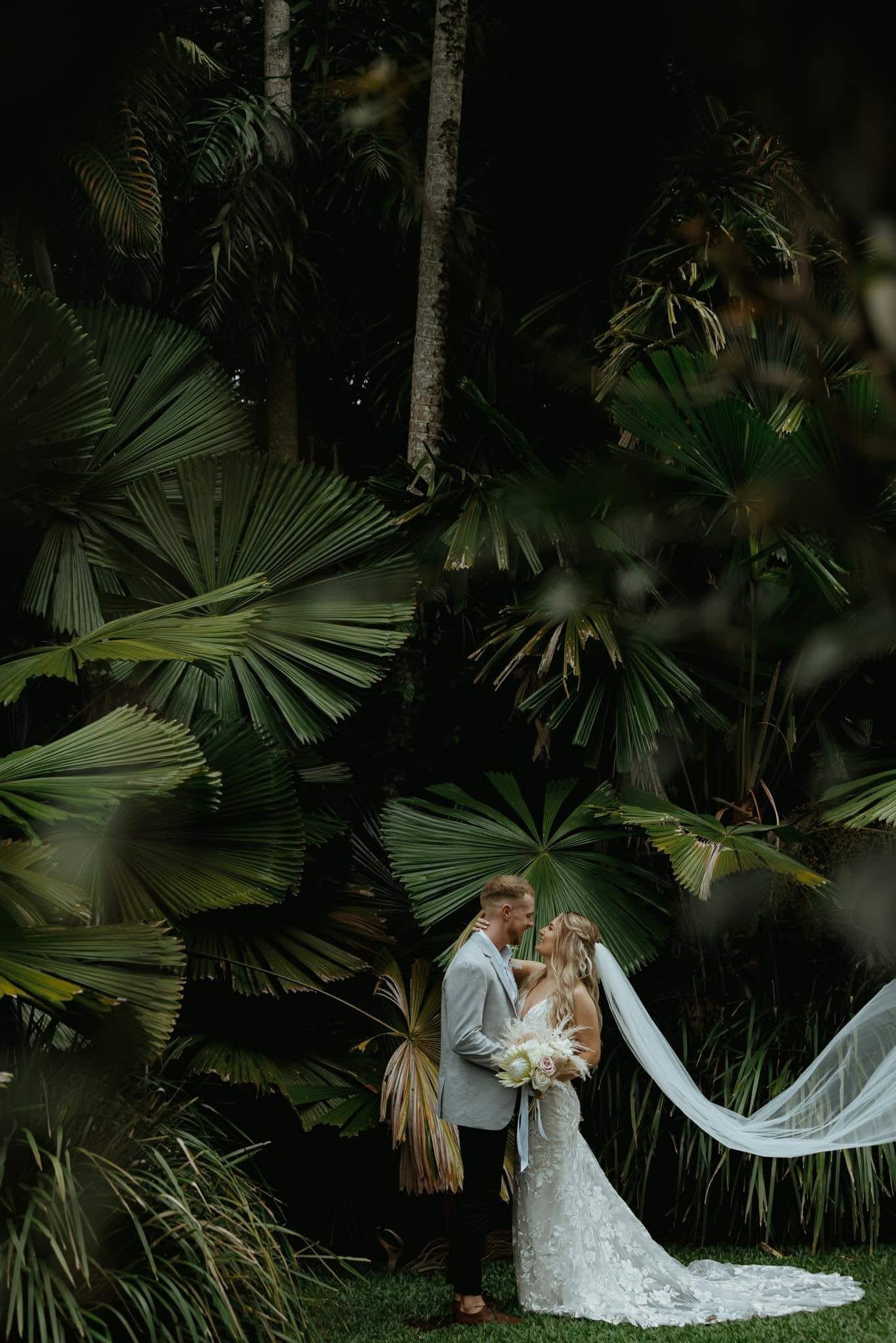 A bride and groom are standing close together in a lush, dark garden filled with large green tropical leaves. The bride is wearing a white lace wedding gown with a long veil and is holding a bouquet of pale pink and white flowers. The groom is dressed in a light gray suit and is gazing at her affectionately.