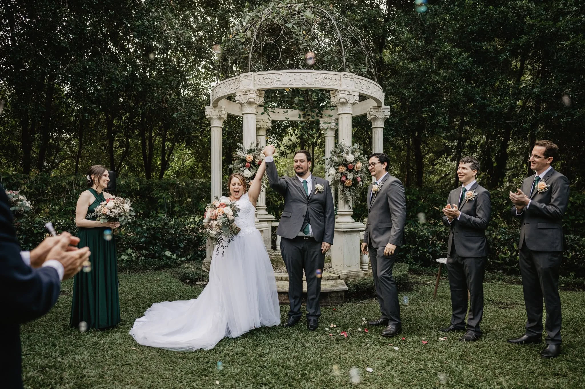 A wedding ceremony outdoors with a bride and groom under a decorative gazebo surrounded by friends and family. The bride is wearing a white dress, and the groom is in a gray suit. Some guests are clapping and celebrating.