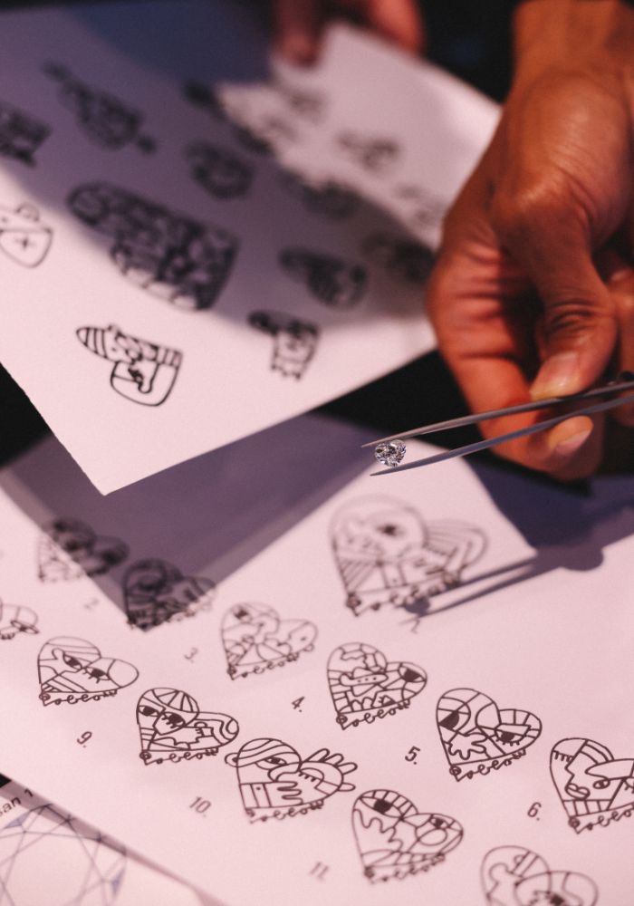 Artist Pablo Lücker using tweezers to hold a small jewel or diamond near coloring pages with heart-shaped stained glass window designs.