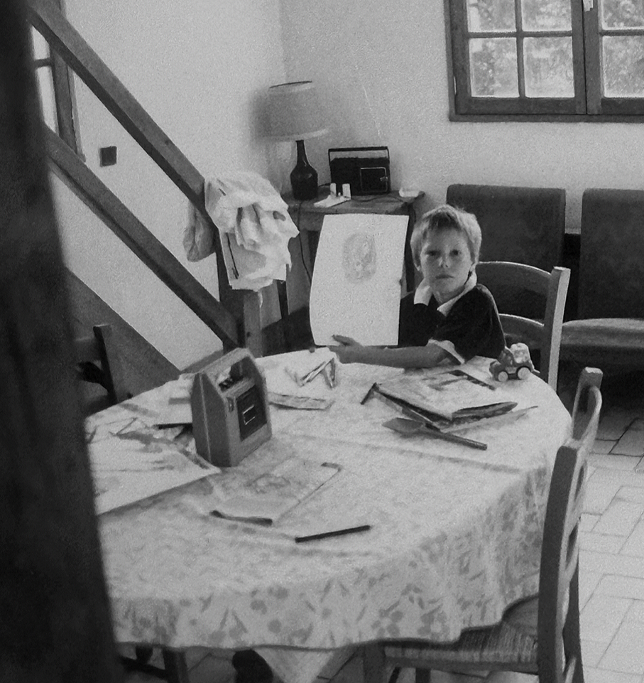 Een jongen zit aan een tafel in een donkere kamer, met tekeningen en kleren op tafel. Hij houdt een tekenbord omhoog en kijkt naar de camera.