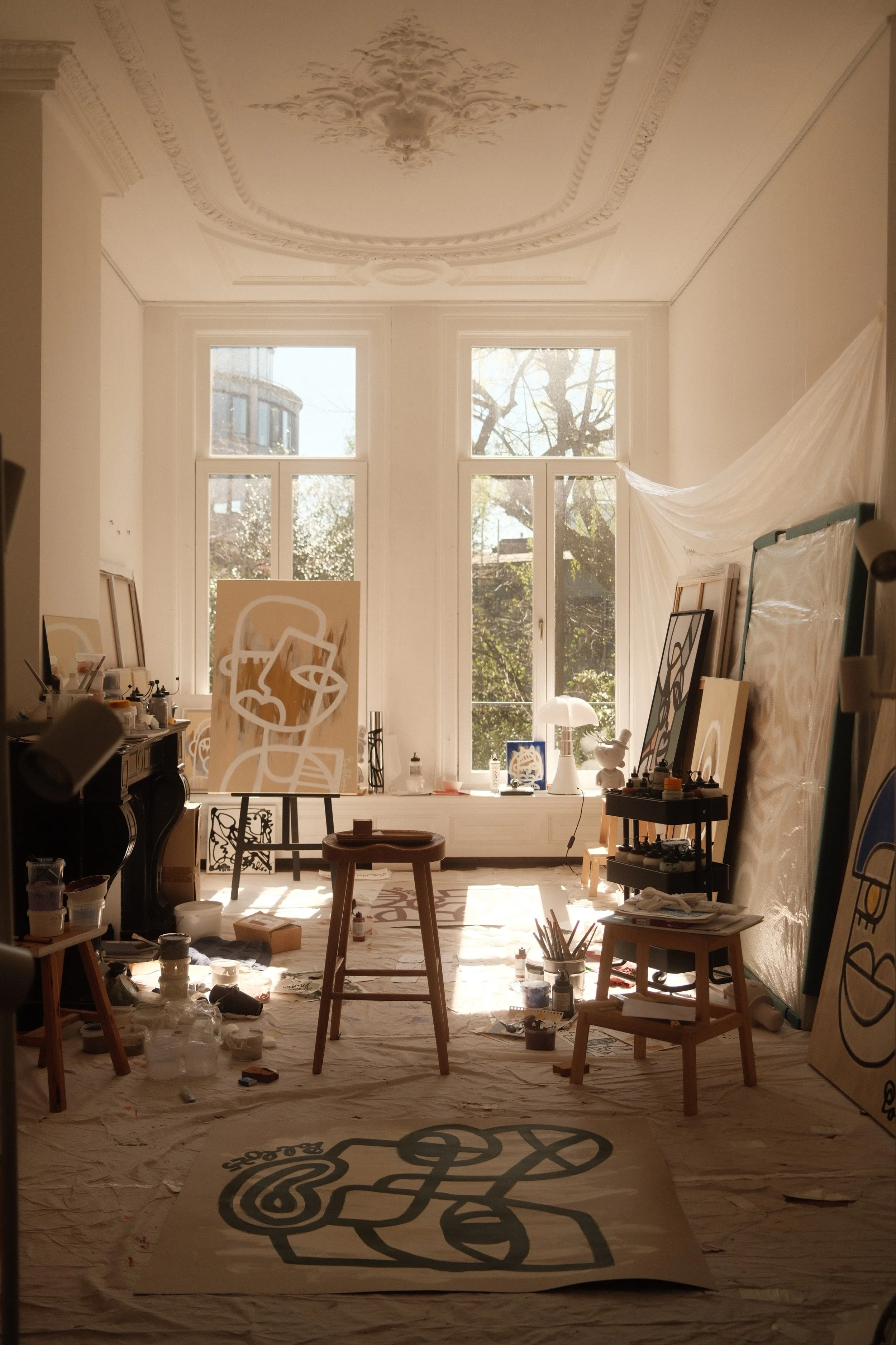 Atelier van Pablo Lucker, contemporary artist, Amsterdam. Een artistiek atelier met grote ramen, waarop kunstwerken zoals portretten en abstracte kunst worden gemaakt, omgeven door schildersbenodigdheden en verlichte door natuurlijk daglicht.