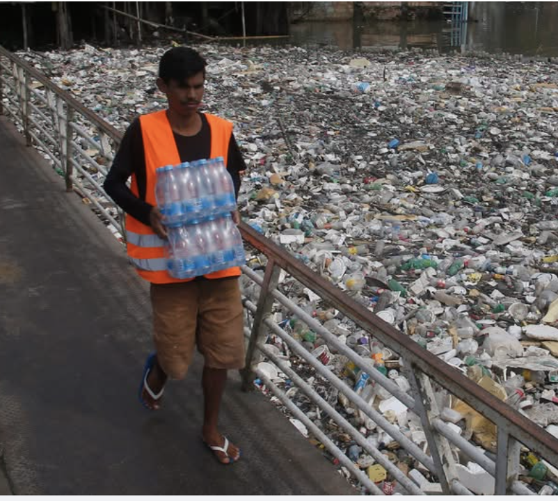 Man in orange vest carrying a stack of plastic water bottles. He's on a walkway. Below the walkway is plastic trash as far as the eye can see.